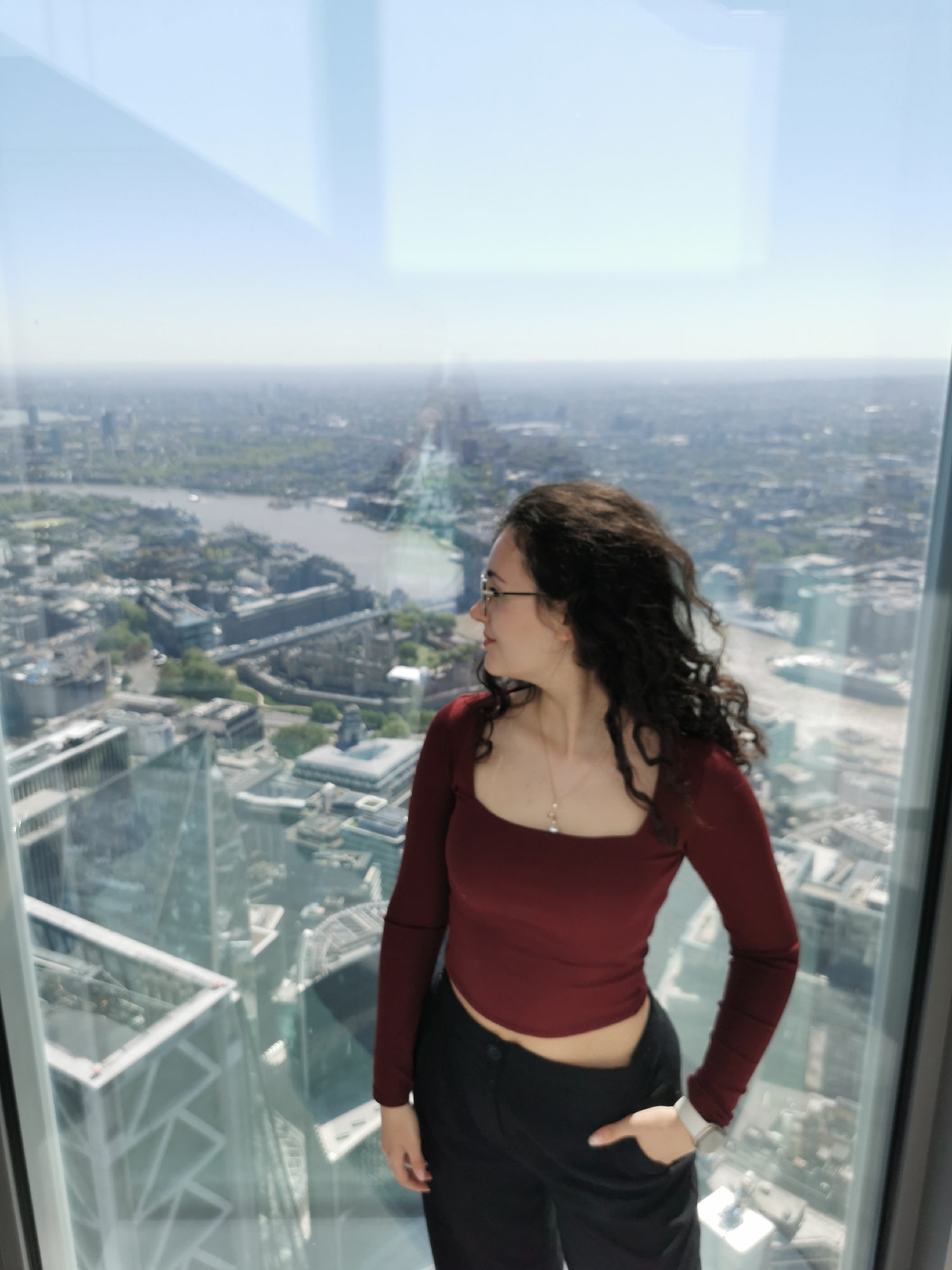 A young woman with dark, curly hair and glasses stands facing to her right, looking out of a large glass window. She wears a dark red, long-sleeved, square-neck crop top and black trousers, with her left hand casually in her pocket. A gold necklace with a small pendant and a silver watch on her left wrist are visible.
The scene depicts her inside a tall building, gazing over an expansive urban landscape. The city, identified as London, is spread out below, with the River Thames clearly visible winding through it. Numerous buildings, bridges, and green spaces dot the dense cityscape, extending to the horizon under a bright, clear sky. Light reflections of internal structures and the exterior of the building she is in are noticeable on the large glass pane, particularly in the upper and lower left sections, suggesting a modern high-rise structure like a viewing platform.
The time of day is sunny and bright, indicative of midday or early afternoon, with excellent visibility. Her posture and gaze suggest a moment of contemplation or appreciation of the vast view. No visible text is present in the image.