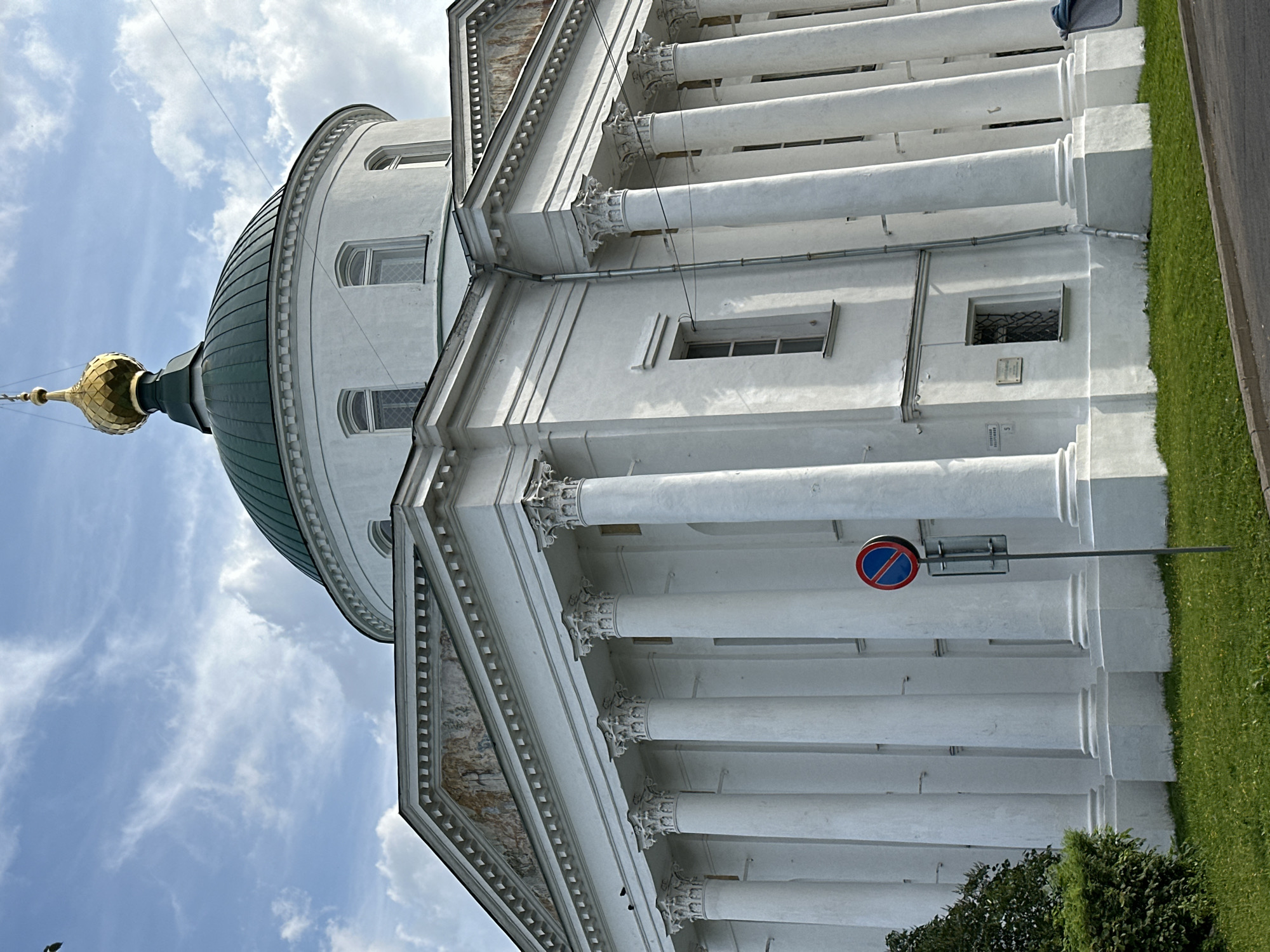 The image shows the white stone exterior of the Holy Trinity Cathedral in Zhukovo, Russia. The cathedral features a green dome topped with a golden cupola. A colonnade of tall white columns supports the building's upper stories. The sky is partly cloudy. A no-parking sign stands in front of the columns. Green grass and a paved road are visible in the foreground.