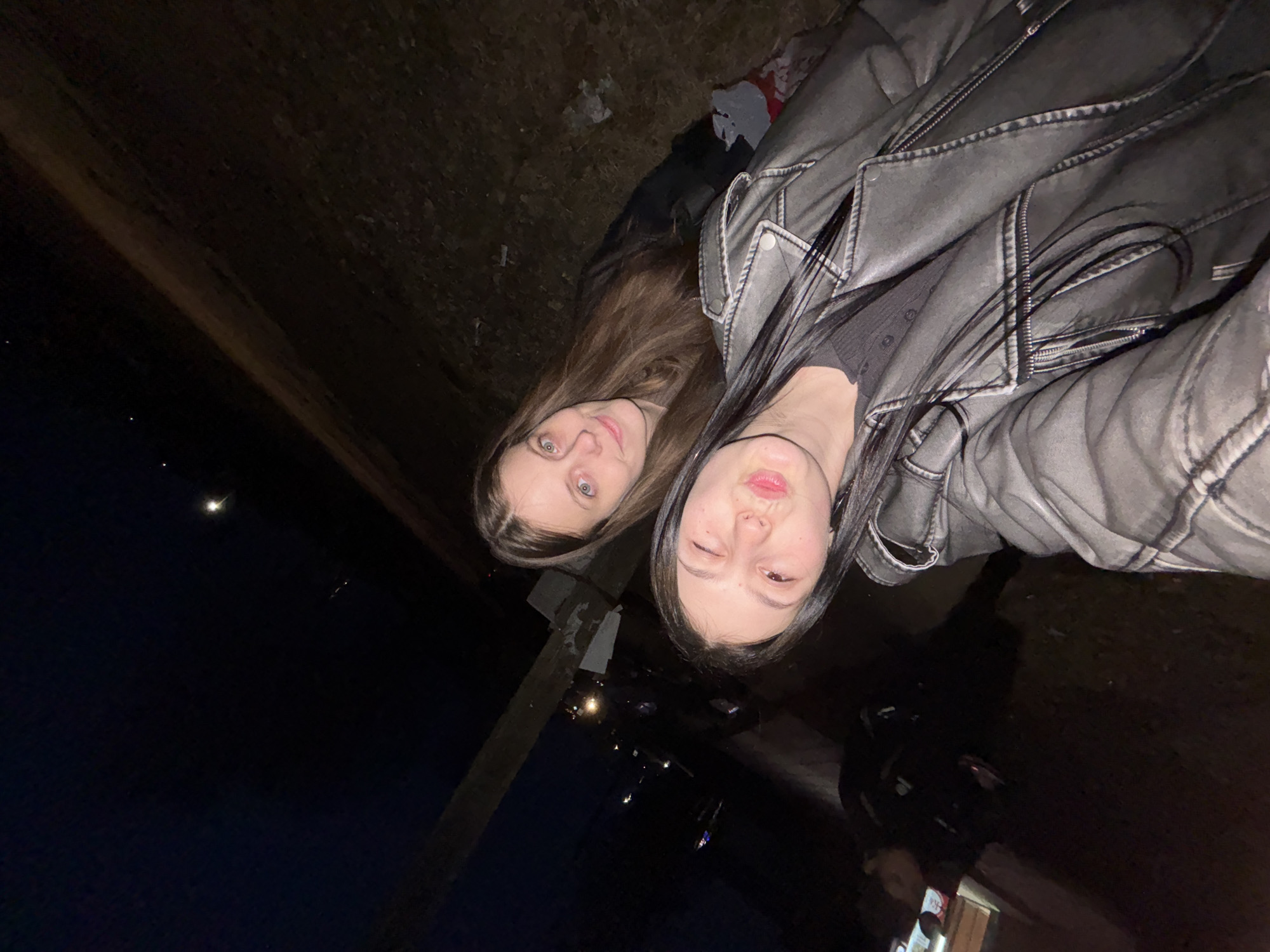 Two young women pose closely for a selfie at night. The woman on the left has long brown hair and blue eyes. The woman on the right has long black hair and is wearing a dark gray leather jacket. The background suggests an urban environment with streetlights and parked cars. A utility pole is visible in the middle of the frame. The photo appears to be taken at ground level. The location is possibly Фоки, Russia, though no definitive landmarks can be determined.