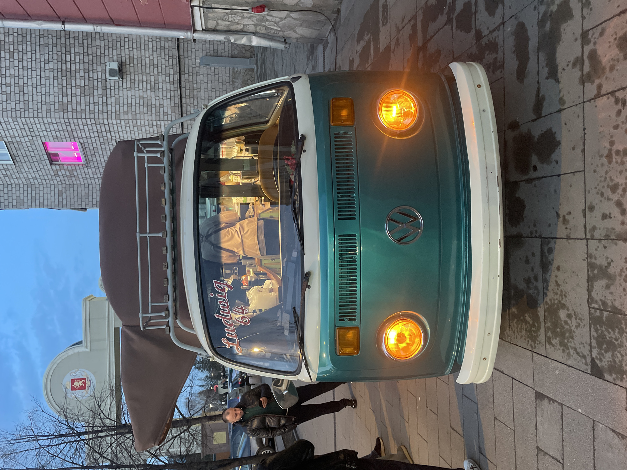 The image shows a teal and white Volkswagen van with a roof rack parked on a wet stone-tiled sidewalk. The van has the name "Ludwig '64" in cursive on the windshield. The van's headlights are on. Two individuals are partially visible near the van, with one appearing to be using a mobile phone. Bare trees are visible in the background. Beyond the trees, a building with a red and white emblem is visible. A building with a pink-lit window is also visible in the background. Based on the architectural style and the emblem on the building, the location is identified as Krasnoyarsk, Russia.