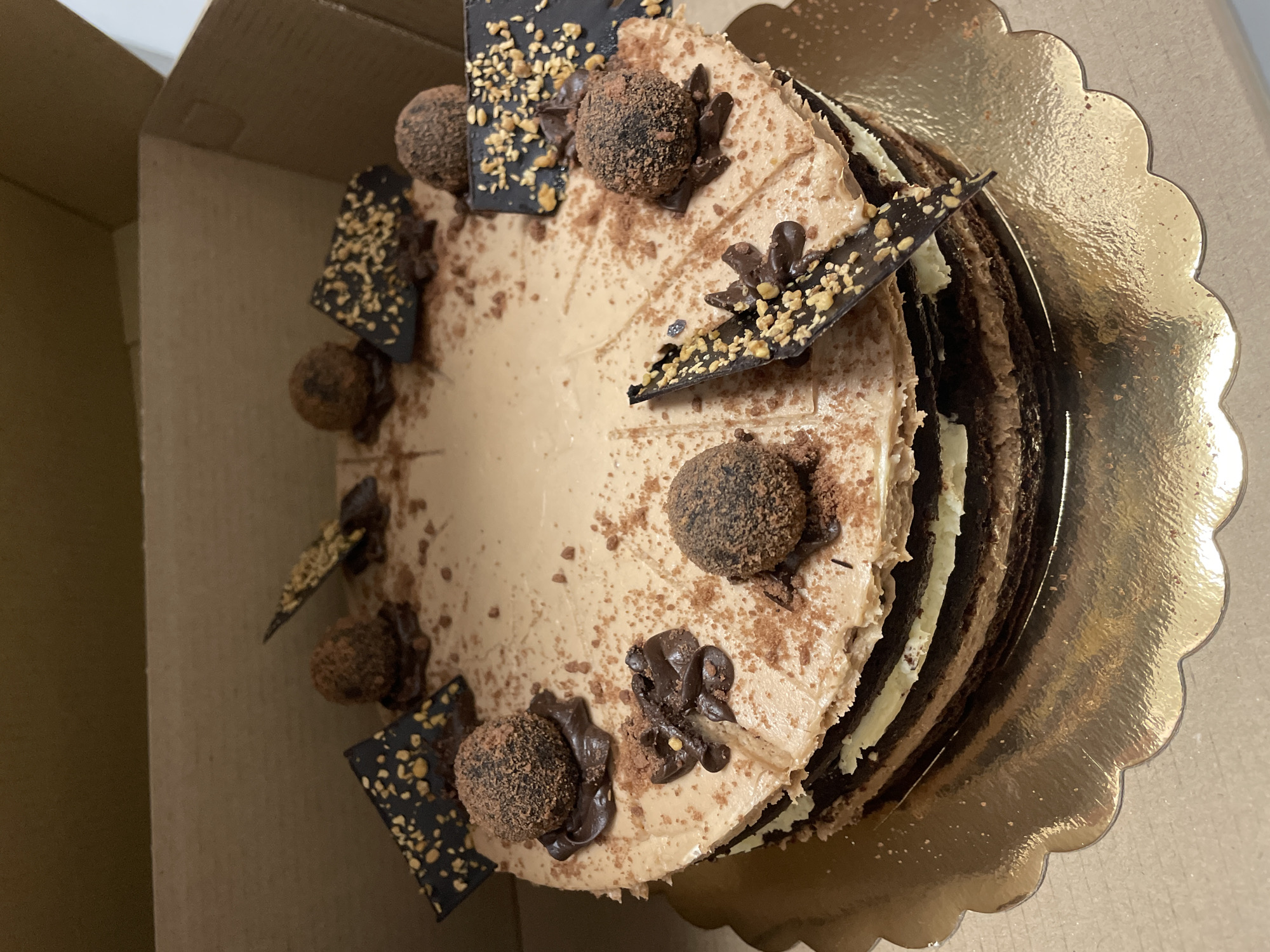 The image shows a chocolate cake in a cardboard box, sitting on a gold scalloped cake board. The cake is decorated with chocolate truffles dusted with cocoa powder, chocolate squares with chopped nuts, and chocolate swirls. The cake appears to have multiple layers of chocolate cake and cream filling. The cake is located indoors in an unspecified location, but given the instruction, is assumed to be in Sofia, Bulgaria.