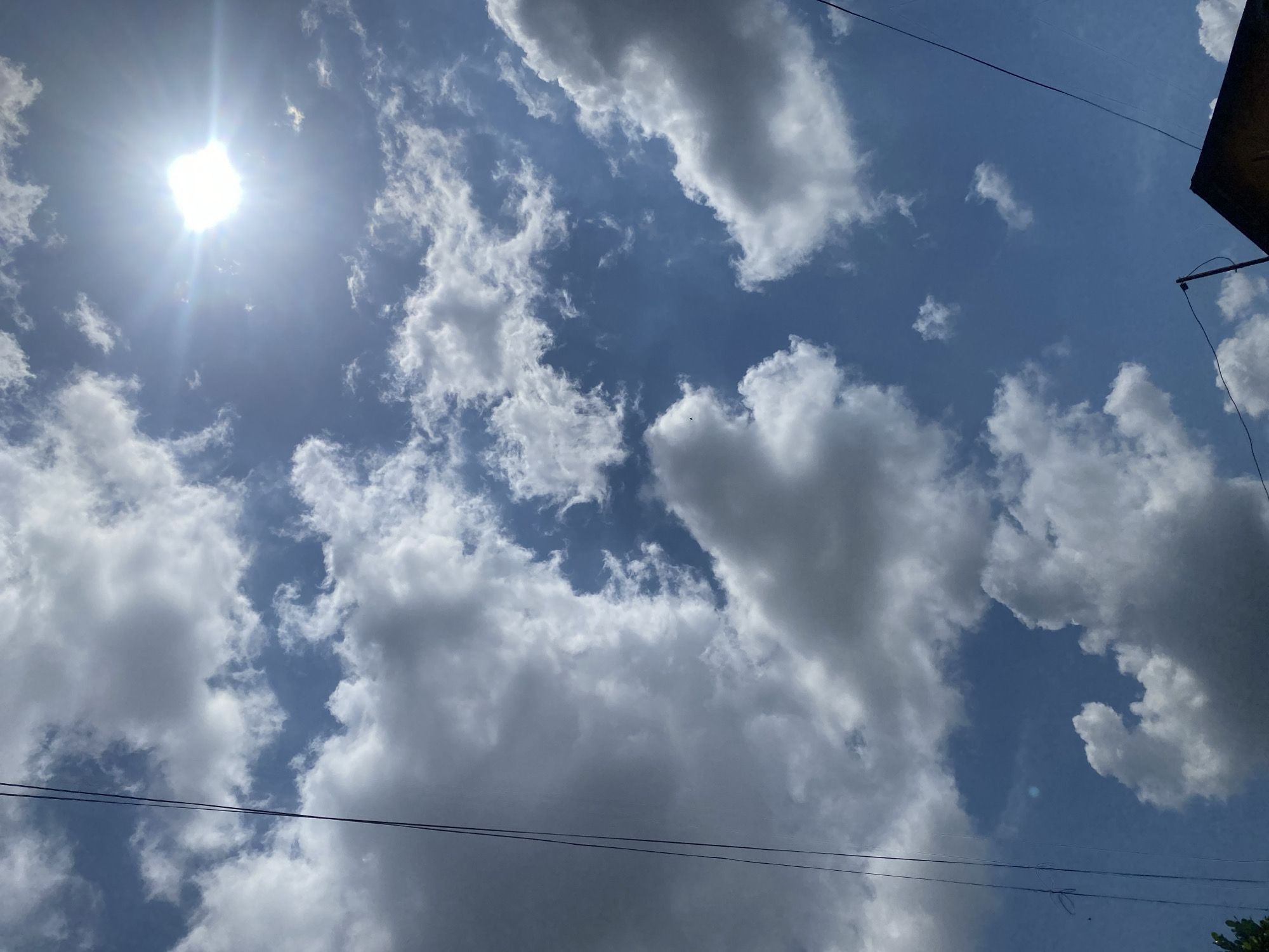 The image captures a bright, sunny day with a clear blue sky filled with fluffy white and gray clouds. The sun is visible in the upper right portion of the frame, emitting a strong glare. Several thin, dark wires, likely power lines or telecommunication cables, are stretched across the sky from the left side and the bottom right. In the bottom right corner, the dark triangular roof of a building is visible, with a pole extending upwards from it. The scene appears to be an outdoor shot looking upwards, possibly taken from a residential area in Berangas Timur, Indonesia, given the presence of utility lines and building structures common in urban or suburban environments. The overall impression is one of a typical day with overhead infrastructure against a backdrop of dynamic cloud formations.
