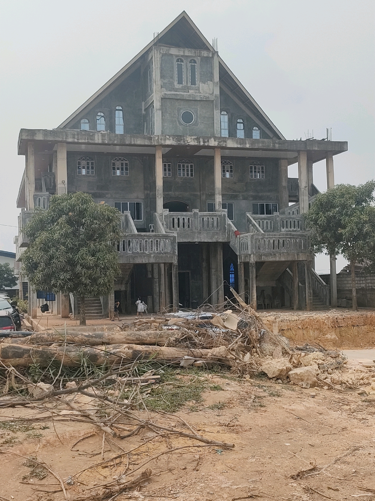 The image depicts a large, unfinished concrete building, possibly a church, with a distinctive triangular roof. The structure has multiple levels, balconies, and numerous windows, some of which are arched. In front of the building, there is a considerable amount of debris, including large tree trunks and branches, as well as rubble and construction materials. A few individuals, including two women in white dresses and at least one man, are visible near the entrance. The ground in the foreground is unpaved, with scattered dirt and grass. The sky is overcast, contributing to a muted color palette. The scene appears to be located in a developing area. There is no identifiable information in the image that places it specifically in Batam, Indonesia.