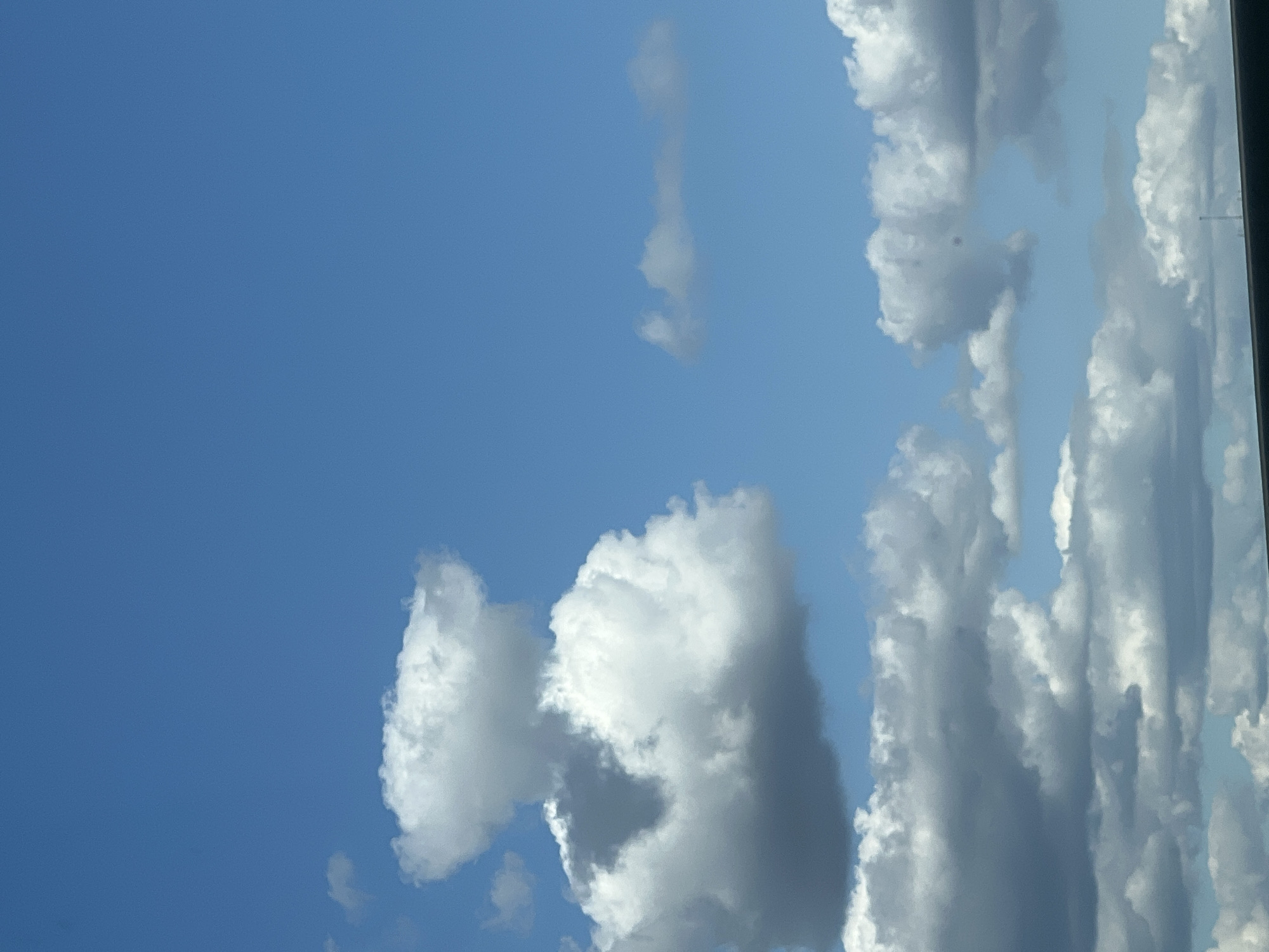 The image displays a bright blue sky with scattered white and gray clouds. The clouds are varied in shape and density, with some appearing fluffy and others more diffused. In the lower portion of the image, a dark horizontal line suggests the presence of a distant structure or horizon. There are no people, animals, or specific landmarks visible in the frame. The scene appears to be an outdoor landscape on a partly cloudy day. The query specifies the location of Warsaw, Poland, but there are no visual cues within the image to confirm or deny this.