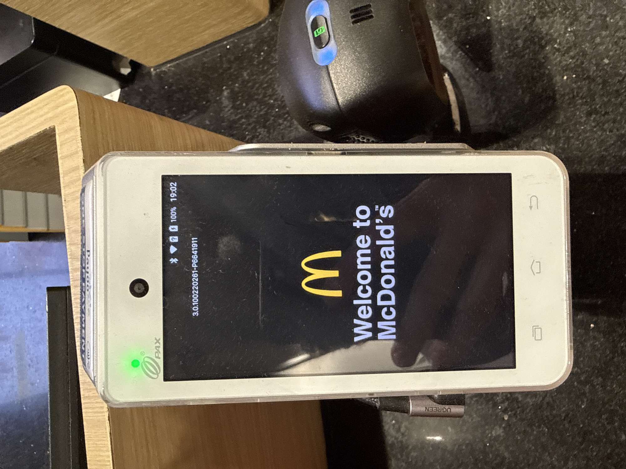 A close-up shot shows a McDonald's branded payment terminal in an indoor setting, likely a restaurant. The terminal is a white, rectangular device with a touchscreen display that prominently features the McDonald's logo and the text "Welcome to McDonald's". A green indicator light is visible above the screen, next to the "PAX" logo, suggesting the device is operational. The screen also displays software version information and the time "19:02".
In the background, a portion of a wooden counter or display is visible, along with a black, textured surface, possibly a countertop. To the right of the payment terminal, a black card reader or similar peripheral device is present, with a blue illuminated section showing a green icon. A USB-C cable, labeled "UGREEN", is plugged into the side of the payment terminal. The overall environment appears to be a commercial establishment, with the lighting suggesting it is a well-lit interior space. The image does not provide specific visual cues that would allow for identification of the city of Nha Trang, Vietnam.