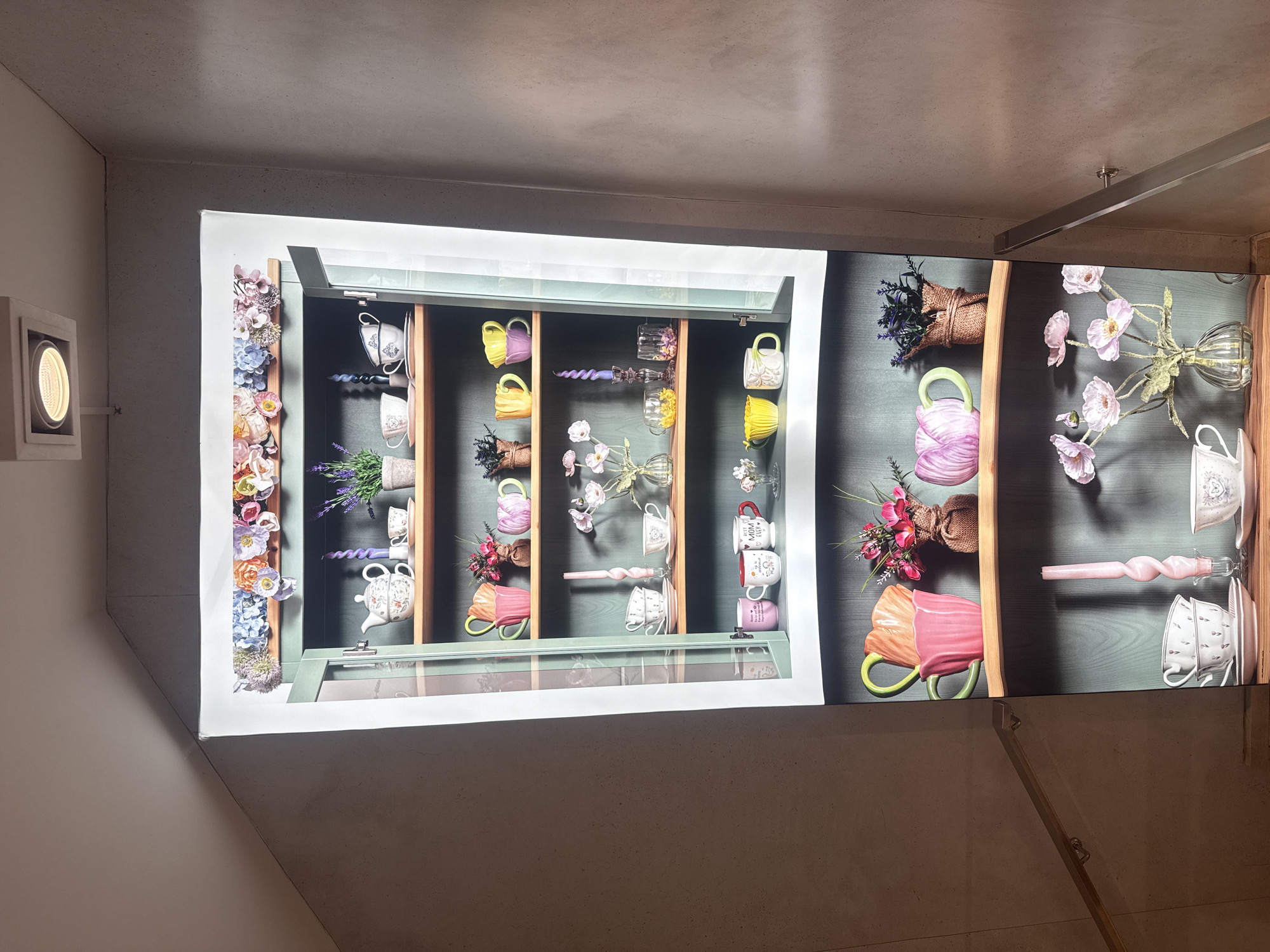 The image displays a large, brightly illuminated vertical panel mounted on a light gray, textured wall, depicting a stylized cabinet filled with an array of decorative items. The digital rendering inside the panel shows multiple shelves holding various teacups, mugs, teapots, and artificial floral arrangements.
The collection includes numerous colorful teacups and mugs in shades of yellow, purple, pink, orange, and white, some whimsically shaped like flower petals. Delicate white teacups and a teapot adorned with intricate patterns are also visible. Artificial flowers such as lavender, pink poppies, and mixed blossoms are arranged in glass vases or rustic woven baskets. Other decorative elements within the cabinet include twisted candles in pink and purple, and small clear glass objects. Notably, one white mug near the right edge of the display is inscribed with the text "BEST MOM EVER <3".
The surrounding environment consists of a plain, possibly concrete or painted plaster wall. To the left of the display, a rectangular recessed light fixture emits a warm, circular glow. A metallic handrail or bar is partially visible in the bottom right corner, suggesting the presence of a staircase or ramp in the immediate vicinity.
The scene is static, featuring no human subjects or activity; its purpose appears to be decorative, creating an aesthetically pleasing focal point. The vibrant colors and floral motifs within the display convey a cheerful and welcoming mood, contrasting with the more minimalist and industrial look of the surrounding interior space. The time of day is indeterminate due to the indoor setting and artificial lighting.