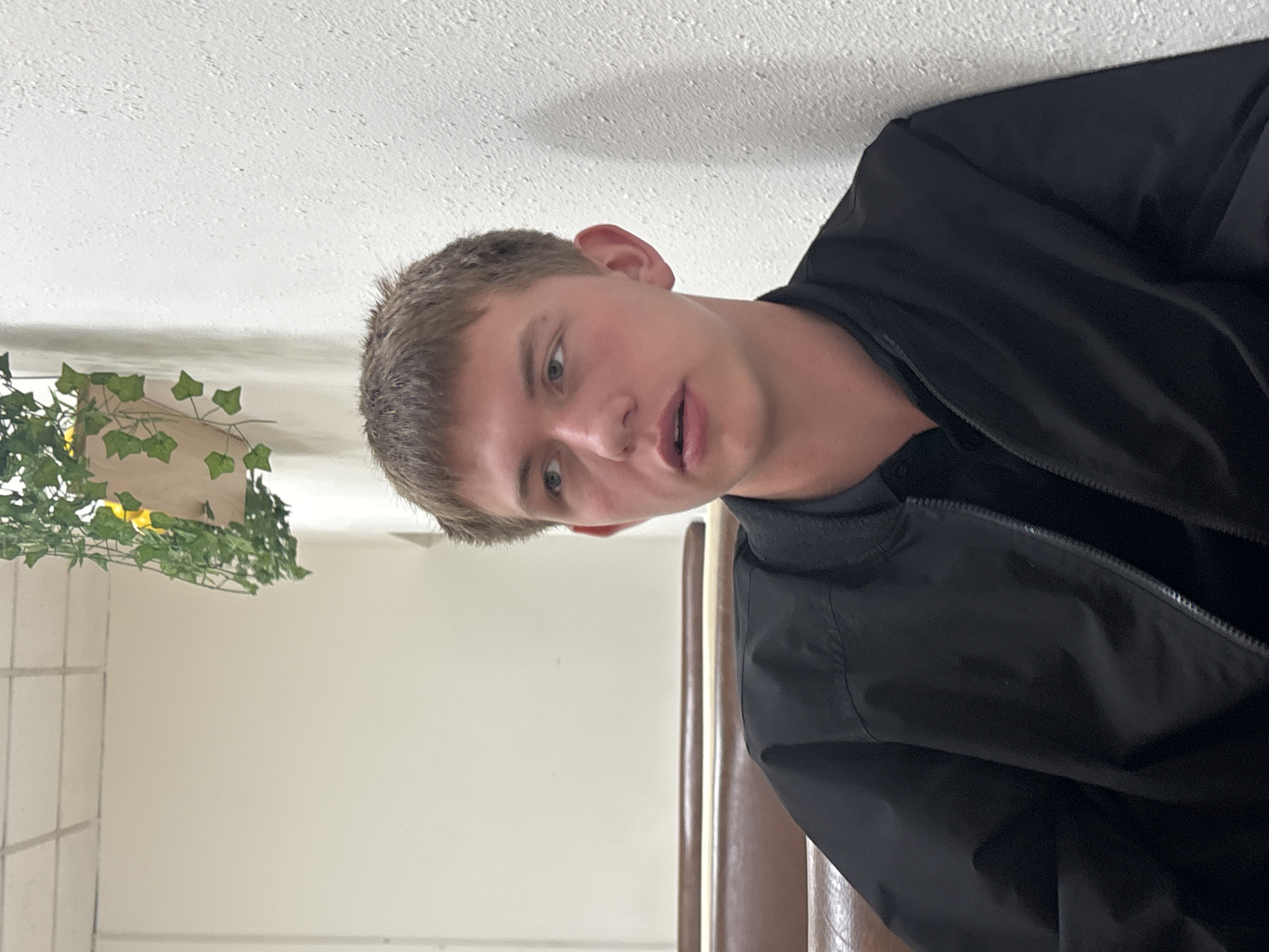 A young male is depicted in a close-up, head-and-shoulders shot within an indoor setting. He has short, light brown hair, light-colored eyes, and fair skin, with his mouth slightly ajar as if speaking or about to speak. He wears a black bomber jacket with a visible zipper over a dark undergarment.
The background shows a textured white ceiling above and to his left. A light-colored wall is visible, against which an indoor plant with green, ivy-like leaves in a light-colored planter is situated. Further to the left, a section of a tiled wall, possibly white or light beige with visible grout lines, is apparent. To the right of the male, a brown, possibly upholstered or wooden, piece of furniture, such as the back of a chair or booth, is partially visible.
The lighting is bright and uniform, suggesting an indoor environment. The combination of ceiling, plain walls, a tiled surface, and casual furniture indicates a public or communal space, such as a cafe, waiting room, or common area. The male's expression is neutral to conversational. There is no visible text in the image.