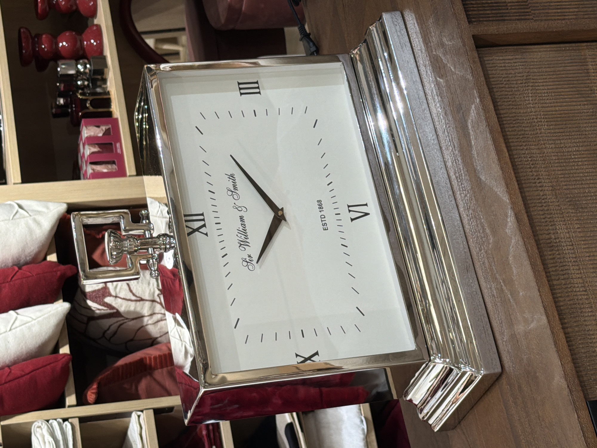 A silver mantle clock with a white face rests on a wooden surface. The clock has Roman numerals for the hours and a brand name that reads "Sir William & Smith ESTD 1868". The clock is positioned in front of a shelf unit filled with pillows and decorative items. The shelf unit is made of light-colored wood and contains several red and white pillows, as well as a few small red decorative objects. In the background, a power cord is visible plugged into a black electrical outlet. The wooden surface in the foreground appears to be a tabletop or counter.