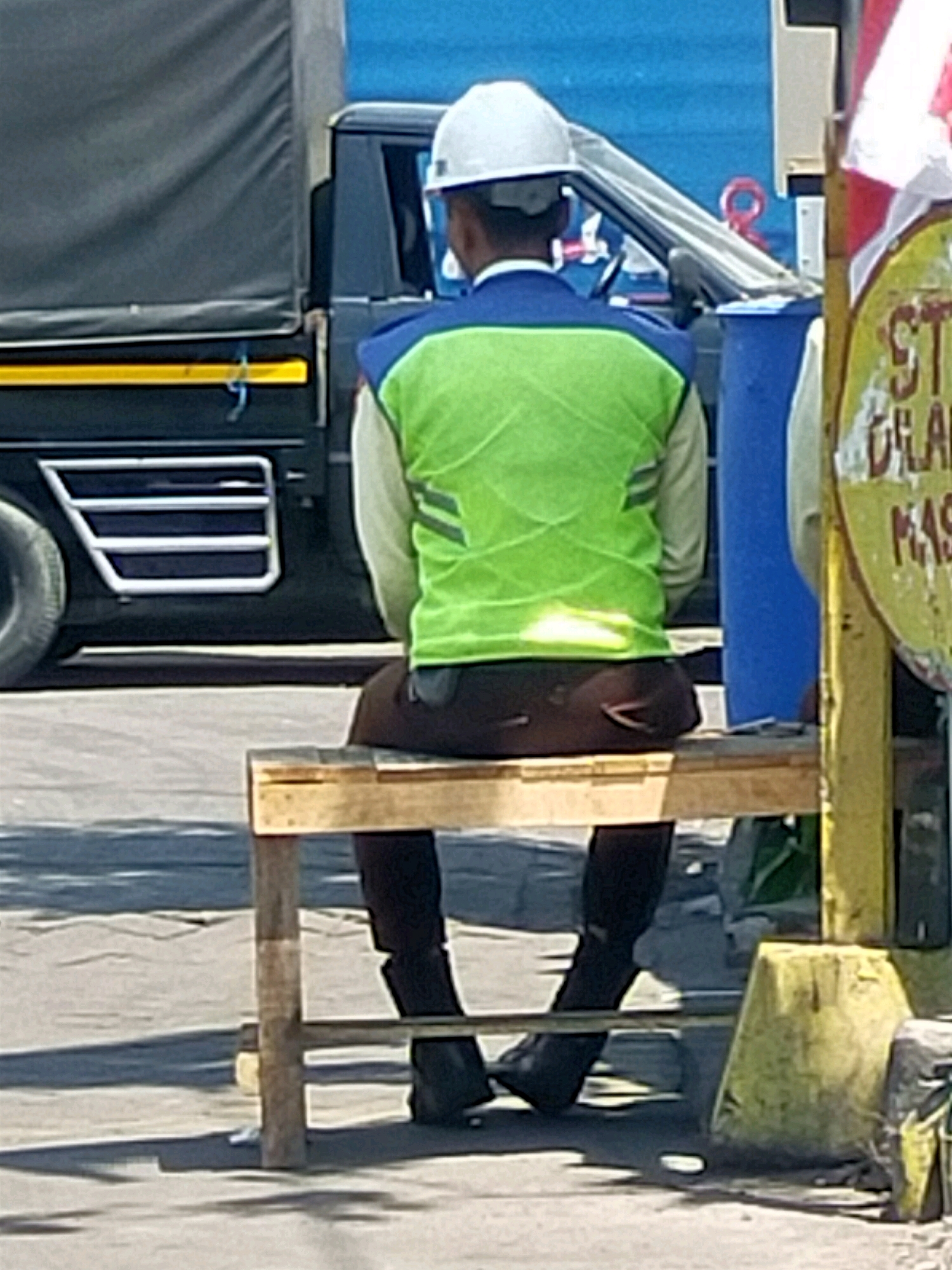 In Surabaya, Indonesia, a person wearing a white hard hat and a bright green safety vest over a light-colored shirt and dark pants sits on a simple wooden bench. They are positioned facing away from the camera. Behind them, a dark-colored truck with a gray canvas cover is visible. To the right of the frame, a yellow pole with a weathered sign, partially obscured by a red and white striped flag, stands prominently. The ground appears to be asphalt or a similar paved surface.
