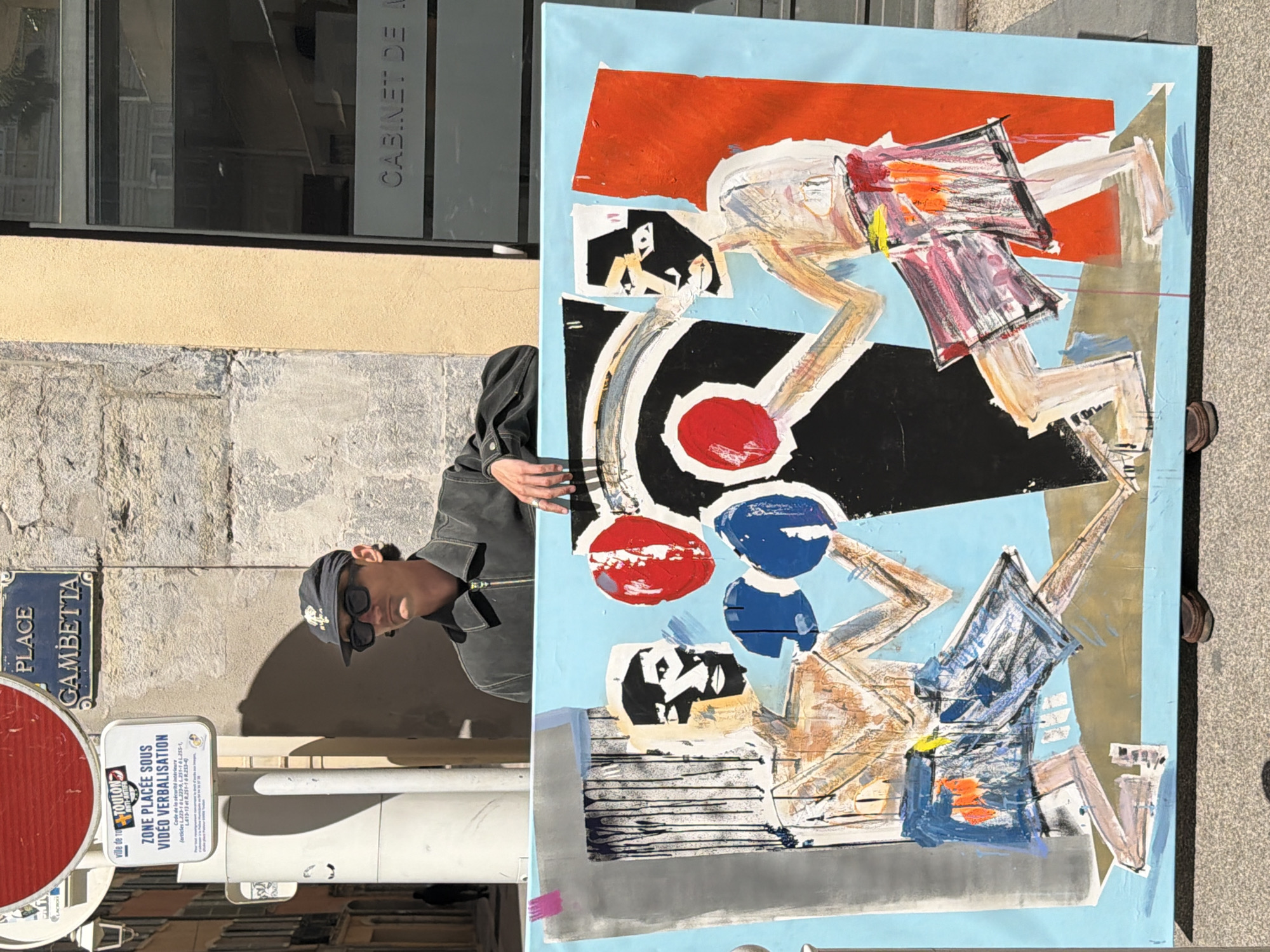Here is a description of the image:
The image shows a person holding a large abstract painting in front of a street scene in Toulon, France. The person, wearing sunglasses and a baseball cap, is partially obscured behind the painting. The painting itself features a light blue background with elements in red, black, white, orange, and shades of gray. The painting appears to depict stylized figures engaged in some sort of action.
In the background, one can see urban elements: a wall, a window labeled "CABINET DE A," street signs, and traffic signals. One street sign reads "PLACE GAMBETTA." Another sign indicates that the area is under video surveillance. The surroundings suggest an urban environment with a mix of older buildings and contemporary signage. The lighting indicates that the photo was taken on a bright day.
