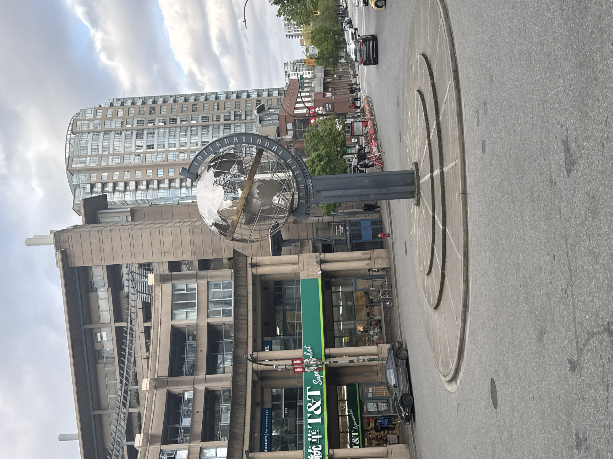 This image captures a bustling urban street scene in Vancouver, Canada, during a cloudy daytime. At the center of the frame stands a large, metallic globe sculpture mounted on a dark pedestal, with the words "INTERNATIONAL VILLAGE" inscribed in an arc above it. The globe depicts a world map.
To the left of the sculpture is a multi-story commercial building made of concrete with numerous windows. The ground floor of this building is home to a T&T Supermarket, prominently indicated by a large green sign displaying "統華T&T Supermarket" in Chinese and English. Other visible text on the building includes "doctors," "optometry," and the number "208." Further back, a modern high-rise building with a curved facade and extensive glass windows rises against the overcast sky.
The asphalt street, visible in the foreground and extending into the background, features a distinct circular pattern paved around the base of the globe sculpture. Several vehicles are present, including a black car parked near the T&T Supermarket and another dark car further down the road. A yellow taxi can be seen in the distance. A few pedestrians are walking on the sidewalks, and a rack of red shared bicycles is parked near a building that has an "OPEN LATE" sign. The sky is uniformly gray with clouds, indicating a diffused daylight condition without direct sunlight.