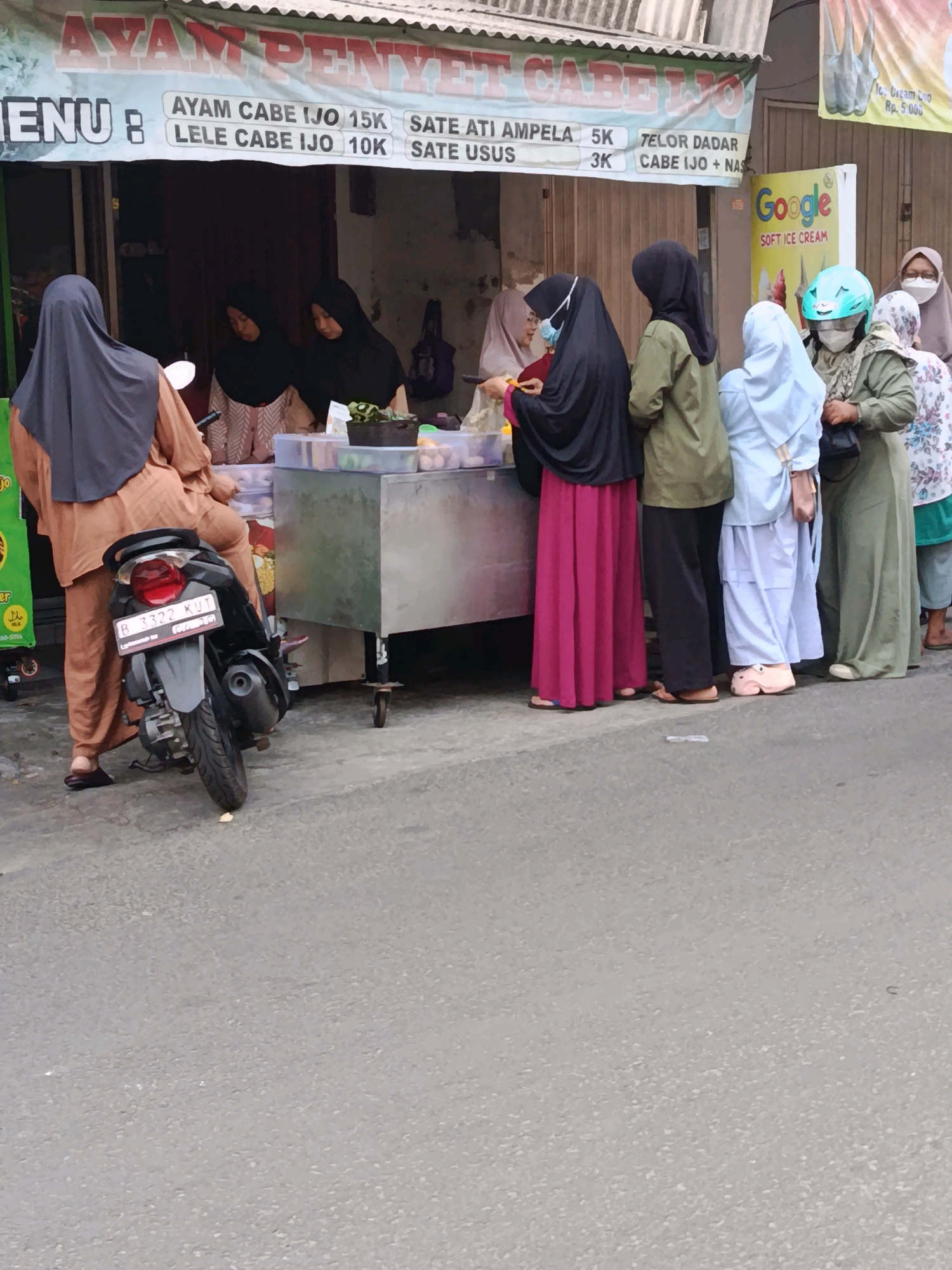 The image depicts a street scene in Bekasi, Indonesia, likely a marketplace or a roadside food stall area. In the foreground, a motorcycle with license plate B 3322 KUT is parked on the asphalt road. A person, partially visible from the back, wearing an orange outfit and a grey hijab, is standing next to the motorcycle.
Behind the motorcycle and the person, a food stall is set up. A large banner above the stall reads "AYAM PENYET CABE IJO" (Spicy Fried Chicken with Green Chili), with a menu list including items like "AYAM CABE IJO 15K," "LELE CABE IJO 10K," "SATE ATI AMPELA 5K," "SATE USUS 3K," and "TELOR DADAR CABE IJO + NASI."
Several people are queuing at the food stall, mostly women wearing hijabs and modest clothing. Some are actively being served or waiting for their orders. The stall itself is a metal cart with containers holding food items. To the right of the stall, a bright yellow sign advertising "Google SOFT ICE CREAM" is visible, featuring images of ice cream cones. Another person wearing a blue helmet and a green outfit is also part of the queue. The overall environment appears to be an outdoor setting with a slightly overcast sky.