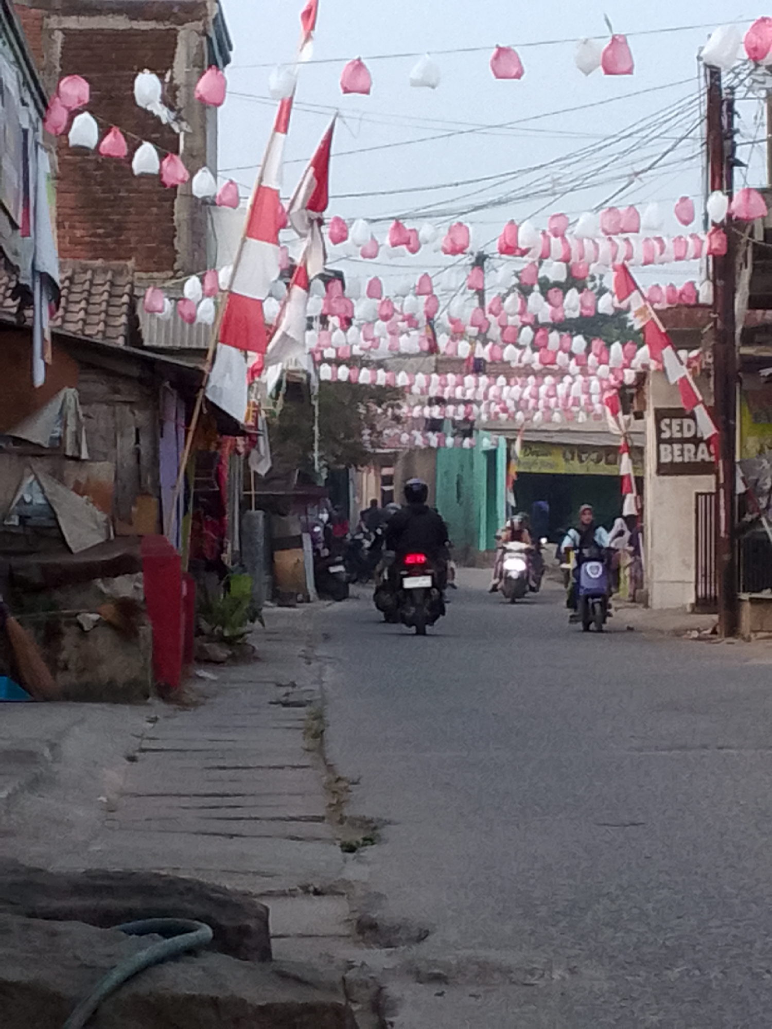 The image captures a street scene in Padalarang, Indonesia, adorned with red and white decorations, possibly for a national celebration. A string of red and white paper lanterns hangs overhead, along with Indonesian flags. Several people are seen riding motorcycles on the paved road. On the left side of the road, there are buildings with visible brickwork and tiled roofs. A sign reading "SED BERA" is visible on one of the buildings. The environment appears to be a moderately populated urban or suburban area. The lighting suggests it could be early morning or late afternoon.