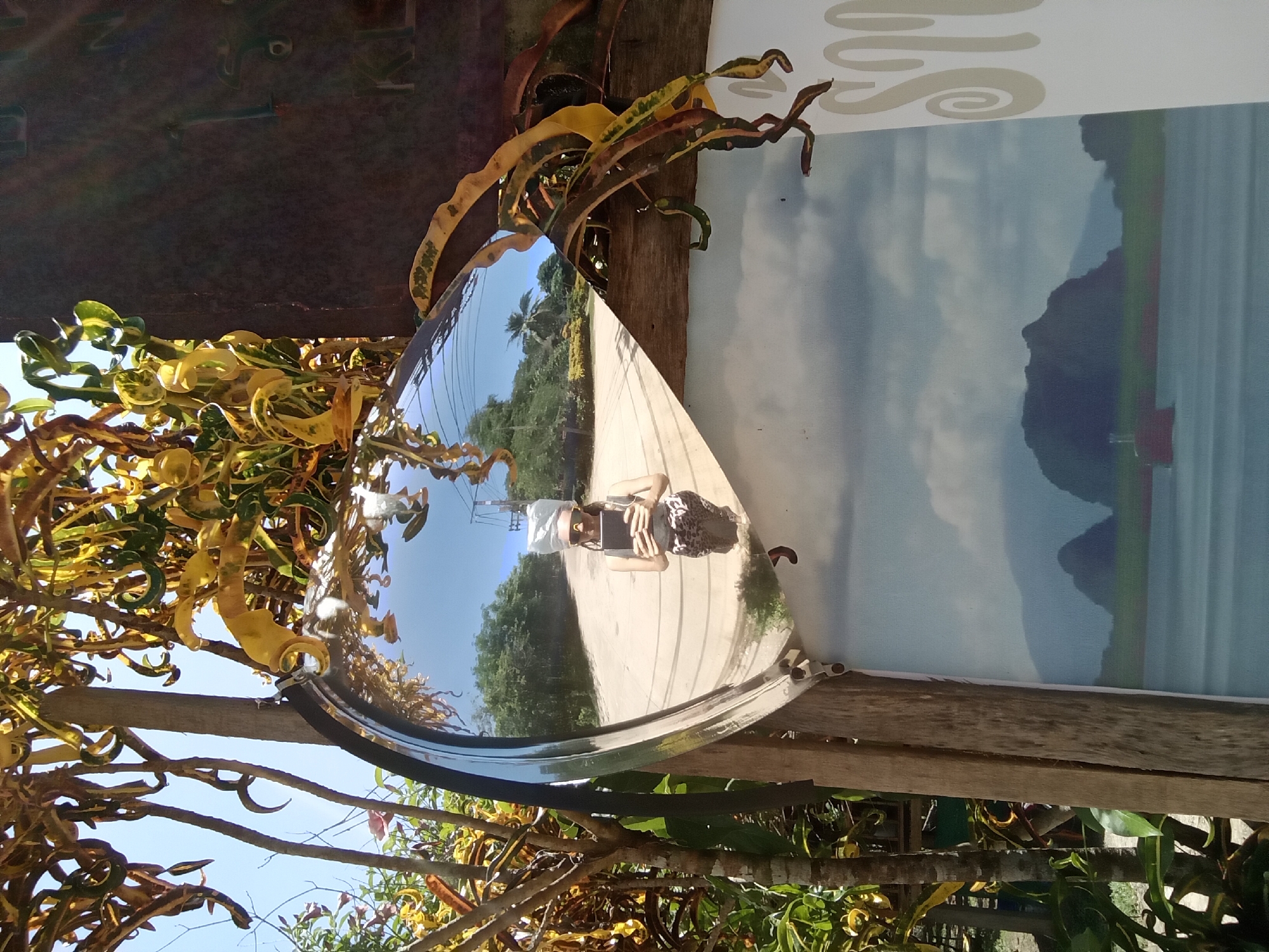 This image depicts a convex security mirror mounted on a weathered wooden frame in an outdoor setting in Ko Siboya, Thailand. The mirror reflects a female individual taking a photograph. She is positioned centrally in the reflection, wearing a light-colored tank top and patterned (possibly leopard print) shorts or a skirt. Her head is wrapped in a light-colored material, and she wears distinctive yellow-framed sunglasses while holding a black electronic device (likely a smartphone or tablet) in front of her. The reflection shows a paved or dirt road receding into the distance, flanked by dense green foliage and trees under a clear blue sky, with visible power lines in the upper right.
Surrounding the mirror, abundant tropical foliage, primarily Croton plants (Codiaeum variegatum) with their characteristic yellow, green, and orange variegated, curled leaves, is prominently visible. The mirror is attached to a vertical wooden post. Below the mirror and to its right, a poster or sign is partially visible. This sign displays a scenic depiction of dark, karstic islands or mountains rising from a body of water under a cloudy sky. A small red object, possibly a buoy or boat, is present in the water in the poster. Partial decorative script, possibly Thai or stylized English, is visible on the far right edge of the poster. The overall scene suggests a roadside or public area on a tropical island.