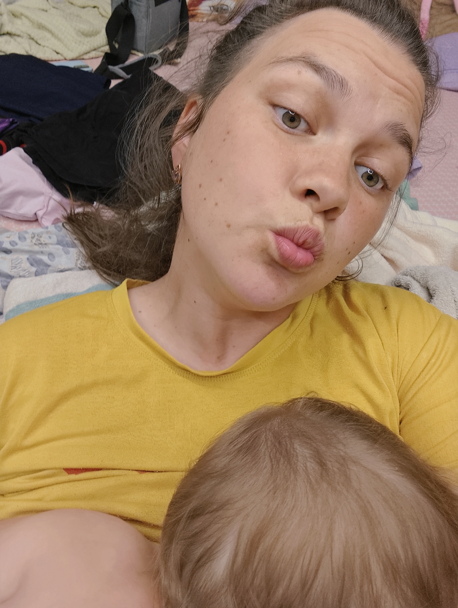 A woman with light skin, hazel eyes, and brown hair pulled back is lying down, making a kissing face. She is wearing a yellow t-shirt. In front of her, resting against her body, is the head of a baby with light brown hair. The background shows a bed with a patterned sheet and various items including clothing and a gray bag. There is no information to definitively determine the location as Chaikovsky, Russia based on this image alone.