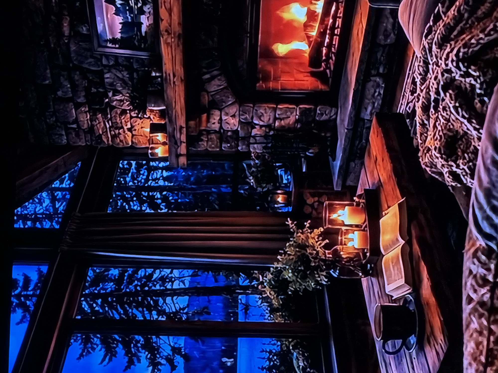 This media file captures a cozy, rustic interior scene, likely a living area within a cabin or lodge, viewed from an elevated perspective looking downwards and to the left. The scene is dominated by warm artificial light contrasted with a cool, dark exterior.
A prominent stone fireplace with a roaring fire is visible in the upper-right portion of the frame, emitting a bright orange glow. Below it, the wall is constructed from rough-hewn stone blocks, extending towards the left. To the left of the fireplace, a large window reveals a dark blue exterior, silhouetting pine or fir trees, indicating it is nighttime or very early morning. Curtains or blinds, drawn partially, frame the windows.
Various rustic details enhance the atmosphere. Several wooden beams and panels are integrated into the structure. On a wooden surface to the right of the window, two wall-mounted sconces hold lit candles, casting a warm light. An open book rests nearby, suggesting an activity of reading. Below the book, a dark-colored mug hangs from a hook. Green foliage, possibly a potted plant or decorative arrangement, is situated near the window and candles. On the far right, a patterned textile, possibly a blanket or part of a cushioned seat, is visible, adding to the comfortable feel. Another framed picture or dark object hangs above the stone wall towards the top-left.
There are no people present in the image, nor any discernible activities beyond the implied relaxation. No visible text can be identified. The overall impression is one of warmth, tranquility, and rustic comfort, perhaps a retreat in a natural setting in Sofiivska Borshchahivka, Ukraine.