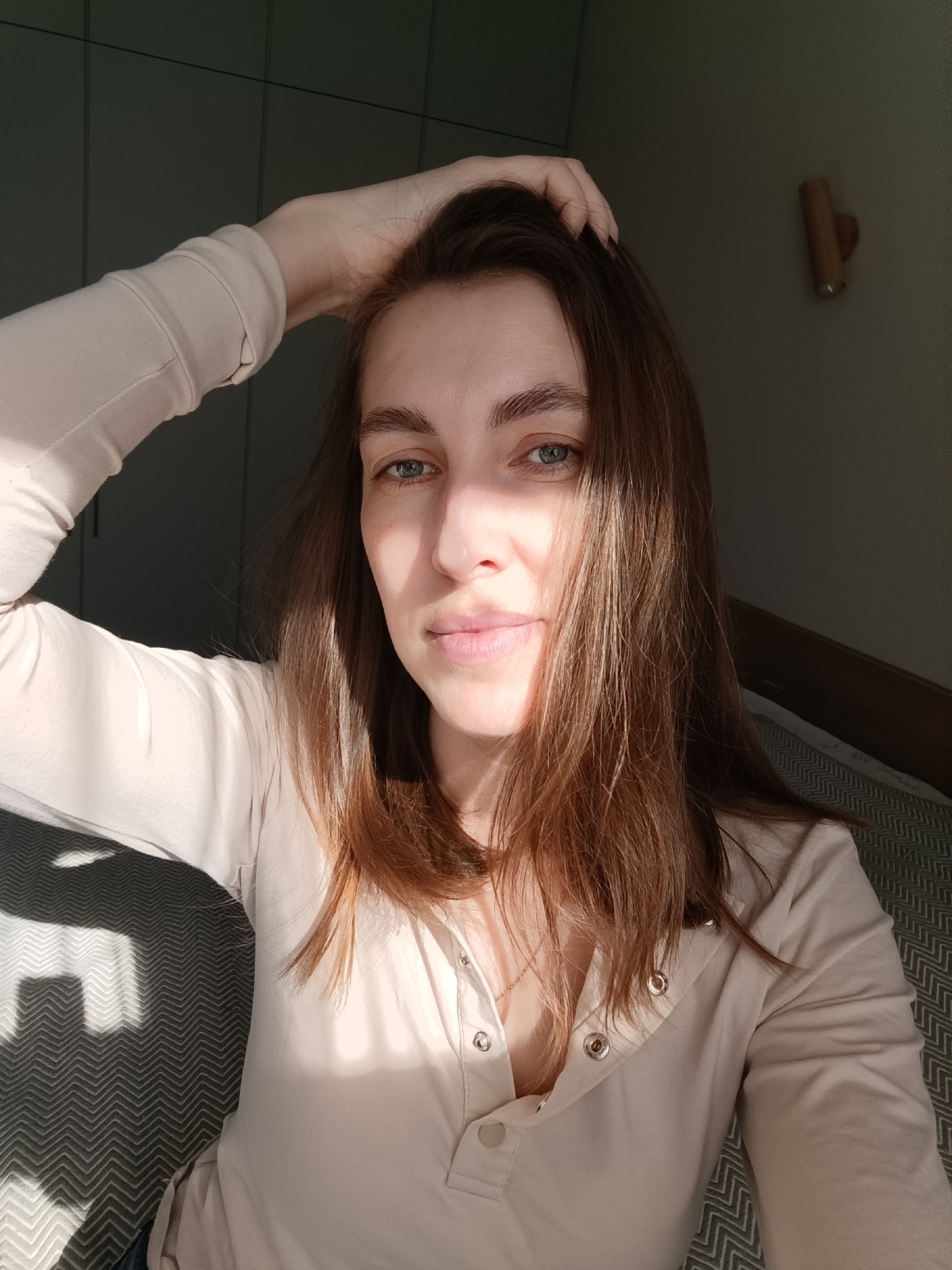 A fair-skinned woman with brown hair and blue eyes is sitting, with her left hand placed atop her head, presumably indoors. She is wearing a light-colored, long-sleeved henley shirt. Sunlight illuminates the left side of her face and hair. The background shows a wall with vertical panels in the upper left corner and a dimly lit section with a wooden rail in the upper right. The seat she is sitting on has a zigzag pattern. The location is speculated to be in Chaikovsky, Russia.