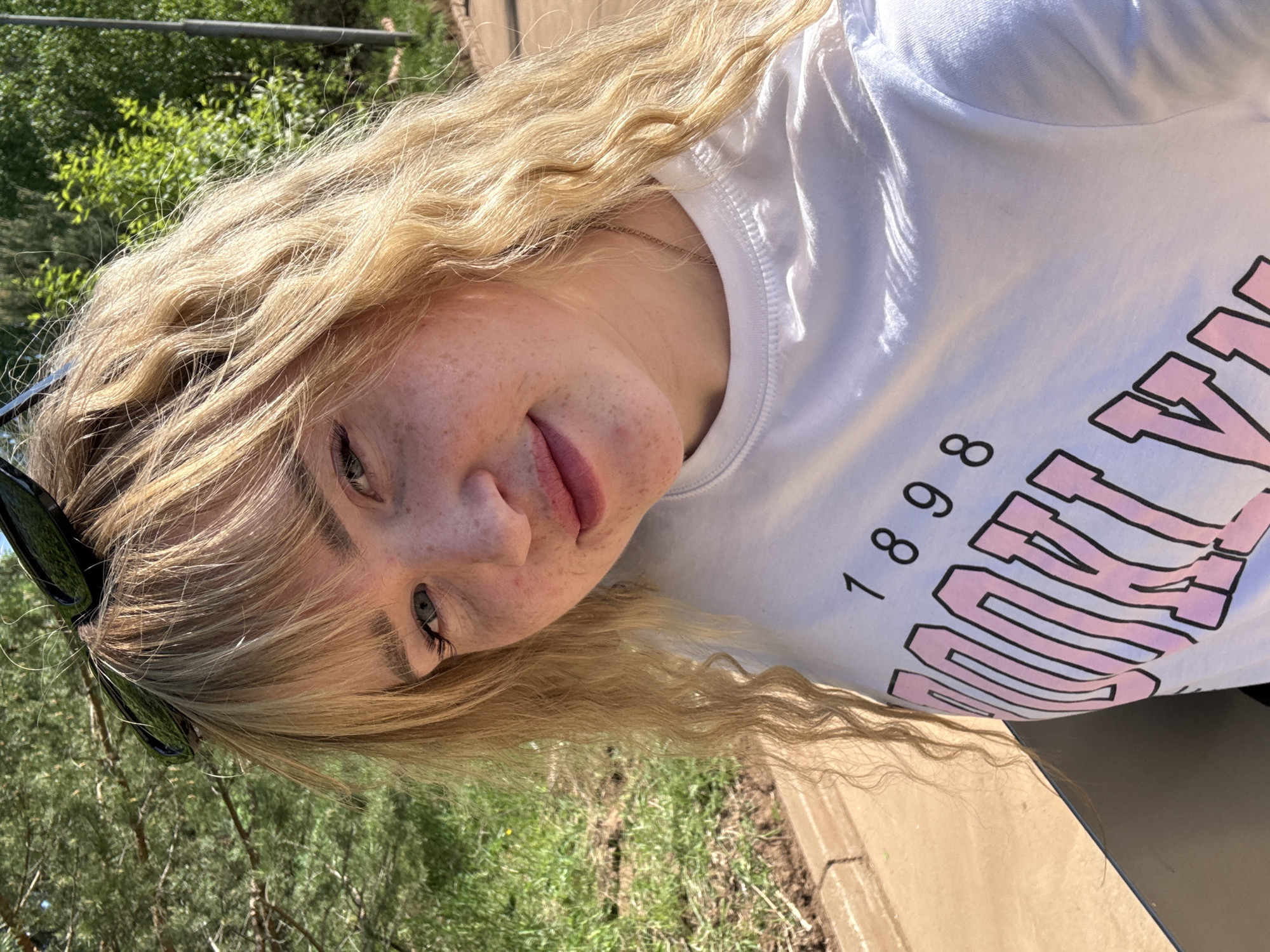 The image shows a woman with blonde wavy hair, wearing a white t-shirt with the number 1898 and the word "SOUTH" in pink lettering, likely outdoors. She has fair skin with freckles. Sunglasses are resting on her head. The background includes green foliage and a wooden or concrete structure. The specific location in Марковский, Russia is undeterminable.