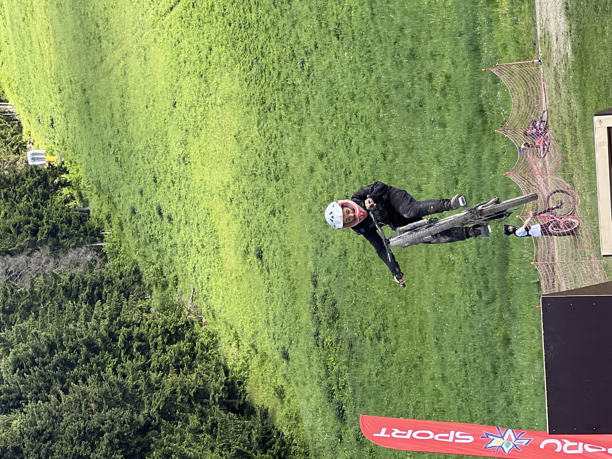 In a natural, outdoor setting, a young person wearing a helmet and protective gear is captured mid-air on a bicycle, performing a jump. The bicyclist is positioned centrally in the frame, leaning forward with the bicycle at an upward angle. Their body is clad in dark clothing, and they wear a white helmet with pink accents. The bicycle appears to be a dirt bike, with knobby tires and a suspension system.
The background consists of a lush, green grassy hill, sparsely dotted with wildflowers. Trees form a dense forest on the left side of the image, with their dark green foliage contrasting with the bright green of the hillside. In the distance, a yellow sign with a black U-shape is visible on the grassy slope.
In the foreground, a red banner with white lettering that reads "SPORT" is planted vertically on the left side. Below the jump area, a dark wooden ramp or platform is visible. To the right of the ramp, a safety net made of orange mesh with thin poles separates the jump zone from the surrounding area. Two other cyclists, one in a white shirt and another lying on the ground with a bicycle, are visible behind the net. The scene appears to be a mountain biking event or practice session.