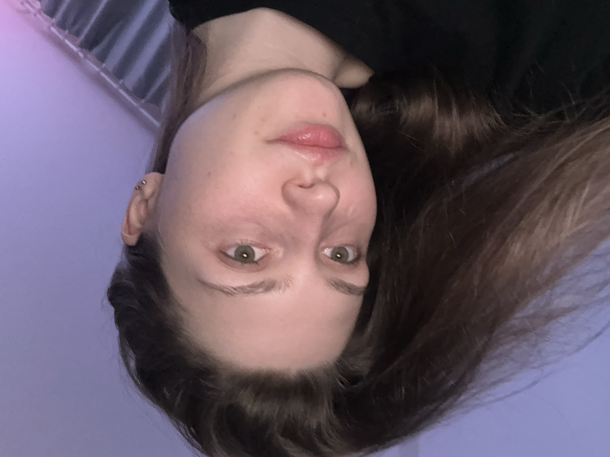 The image shows a young woman's face close-up. She has fair skin, light brown hair, and green eyes. There is a visible piercing on her left ear. The background is a pale purple color with what appears to be a radiator or a similar structure visible at the top of the frame. The image is upside down. There is no information to suggest the location is Saint Petersburg, Russia.