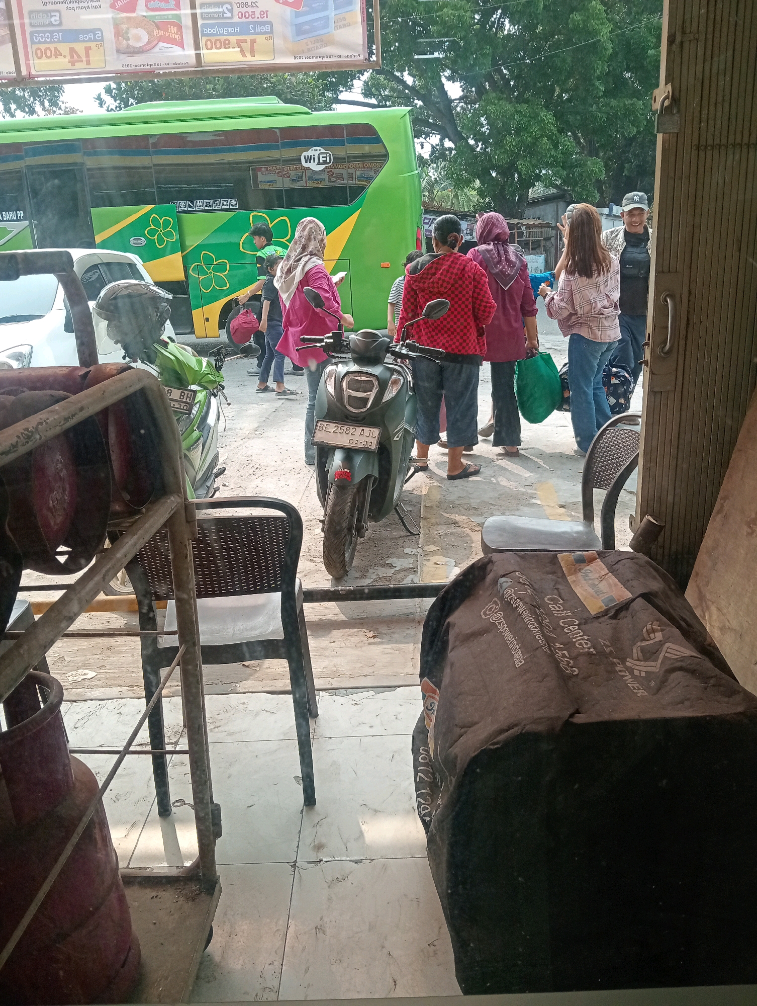 A street scene in Bandar Lampung, Indonesia, shows a green bus with a flower motif parked next to a white car. Several people are standing on the sidewalk, some wearing headscarves. A woman in a red and black checkered jacket carries a red bag. Another woman in a pink hijab holds a green bag. A man wearing a baseball cap smiles. A dark-colored scooter with a license plate ending in "AJL" is parked in the foreground. In the foreground, partially obscured by a metal frame and what appears to be a gas cylinder, are two chairs, one black with a patterned seat and the other empty. A large dark-colored bag with white lettering, including the words "Call Center" and "Fitnes," is draped over a surface in the foreground. The environment appears to be an urban setting with trees visible in the background.