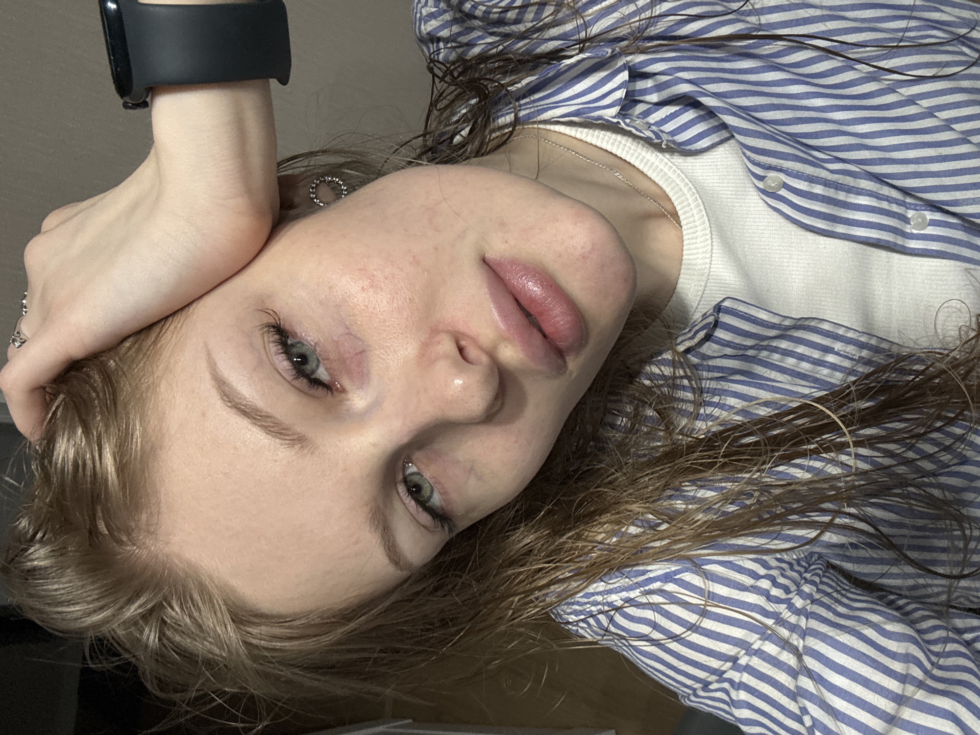 A close-up, angled portrait shows a young woman with light skin, light brown hair (some strands appearing wet), and green eyes. She rests her head on her left hand, where a dark-colored smart watch is visible on her wrist. Her facial features include noticeable pores and some skin texture. She wears a white undershirt beneath a blue and white striped button-down shirt. A thin silver chain necklace is around her neck. A stud earring is in her left ear. The background is a plain, neutral color. There is no discernible indication of the location being Perm, Russia or any other specific city.
