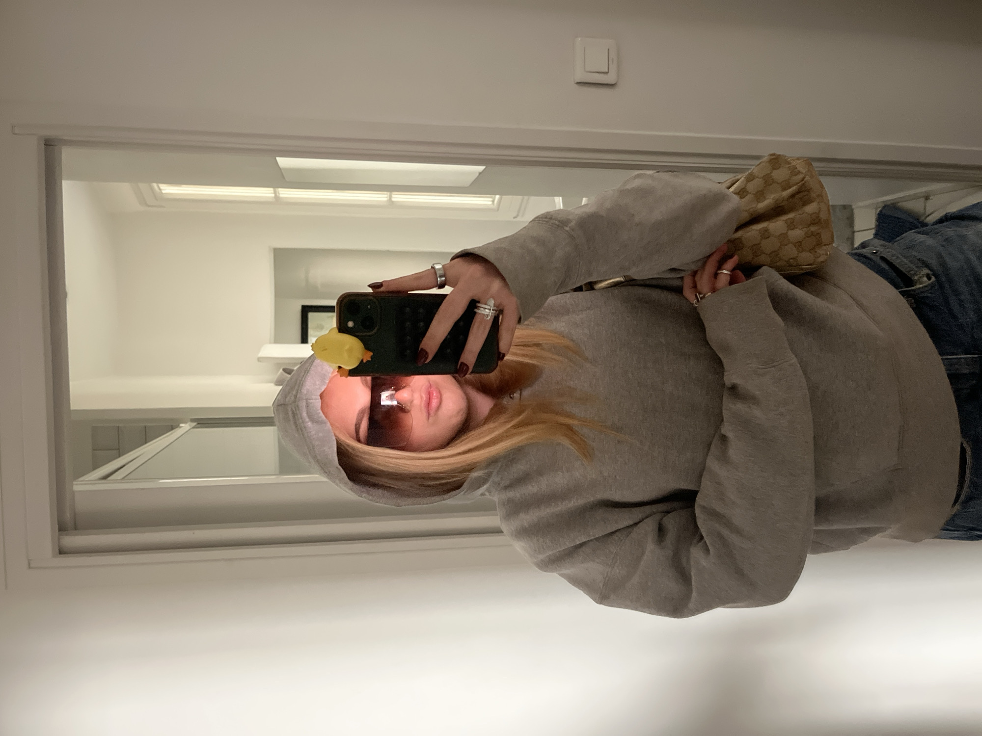 A person wearing a grey hoodie, sunglasses, and jeans is taking a mirror selfie. They are holding a dark-colored smartphone with a yellow object attached. The person is also wearing rings on their fingers. A patterned purse is visible near their shoulder. The background shows a white wall with a light switch and a glimpse of an interior space through the mirror. The location is likely an indoor setting, potentially in an apartment. There are no visible indicators of Paris, France.