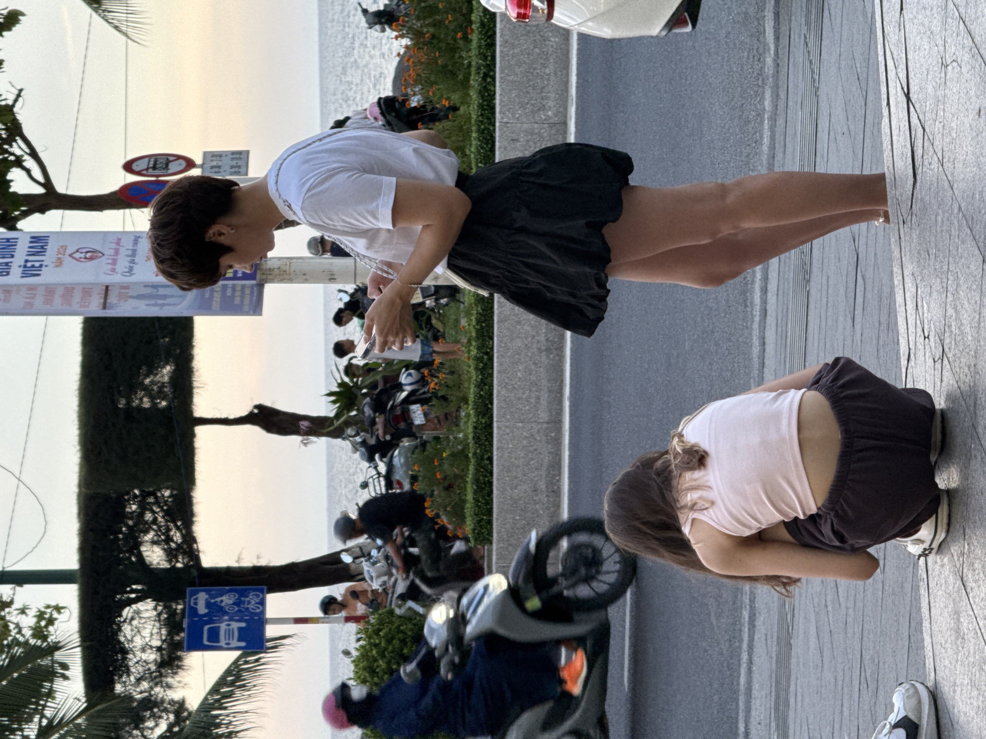 In Nha Trang Ward, Vietnam, a young woman with short hair, wearing a white t-shirt and a black skirt, stands on a paved sidewalk, looking at her smartphone. In the foreground, a younger person with long brown hair, wearing a light pink tank top and dark pants, is crouched down. Behind them, a street is visible with various people on motorbikes, including one rider in a dark blue outfit and a pink helmet. The background shows the ocean with a hazy sky. A street sign with Vietnamese text and a prohibition symbol is present.