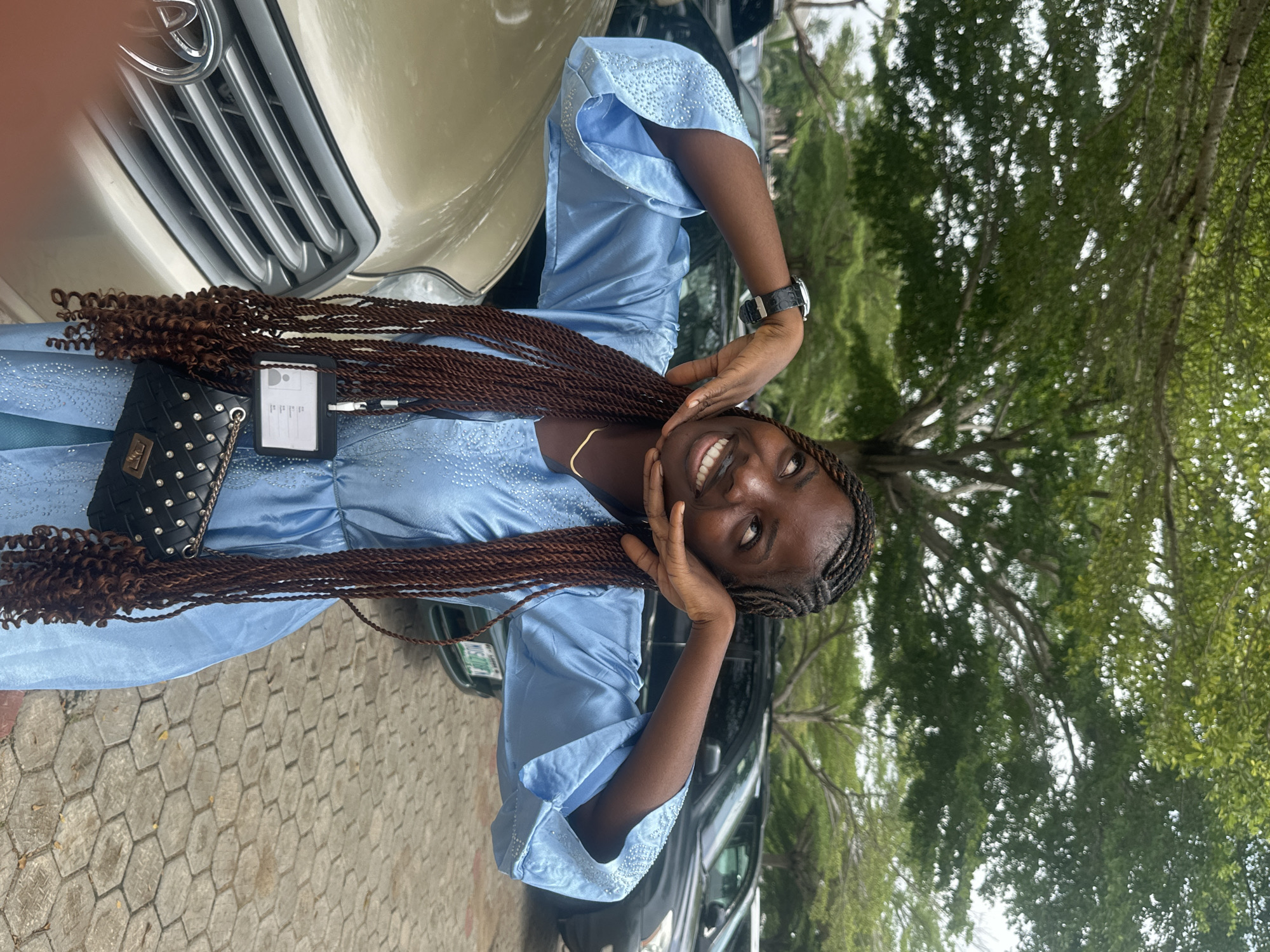 A young Black woman stands outdoors, leaning slightly forward with her hands framing her face. She wears a light blue, long-sleeved dress adorned with small, reflective embellishments on the cuffs and neckline. Her hair is styled in long, dark brown braids that cascade down her front. A black watch with a leather strap is visible on her left wrist. Around her neck, she wears a thin gold chain. A lanyard with a black holder and an ID card is attached to her dress, hanging down near her chest. A small, quilted black handbag with silver hardware is slung over her right shoulder, resting against her hip.
The woman is positioned in front of a light gold vehicle, with its grill and part of the hood visible on the left side of the frame. Behind her, several other vehicles, including dark-colored cars, are parked. The ground is paved with interlocking hexagonal stones. Overhead, the branches of a large tree with green foliage create a canopy, dappled with sunlight. The overall environment suggests an outdoor parking area, possibly in a semi-urban or suburban setting in Itoga, Nigeria. The lighting indicates it is daytime.