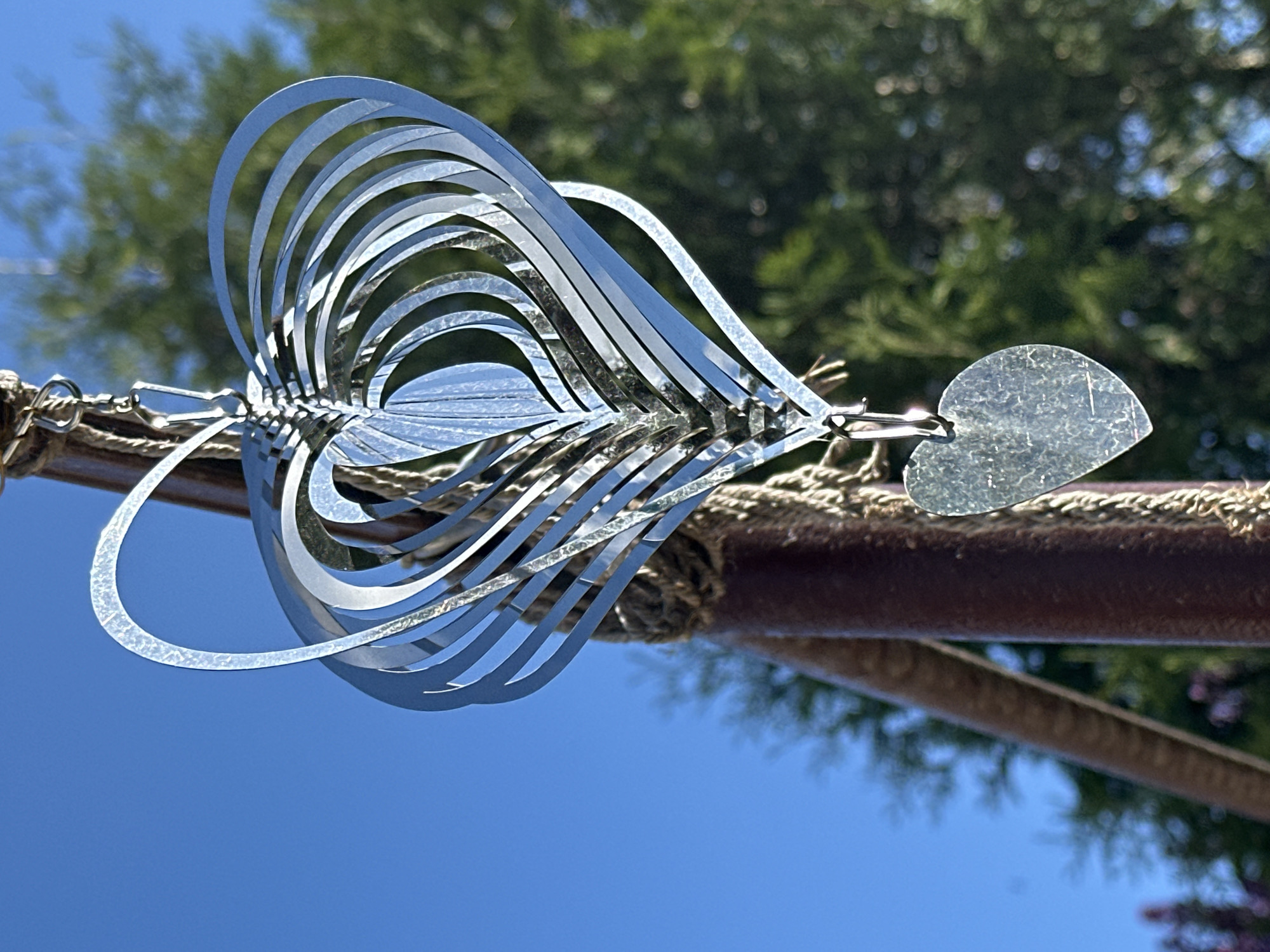 The image shows a metal wind spinner hanging from a rope tied to a metal pole. The spinner is composed of multiple curved metal strips arranged in a heart-like shape. A small, heart-shaped metal piece hangs from the end of the spinner. The background includes a blue sky and blurred green trees. The location is not Minsk, Belarus.