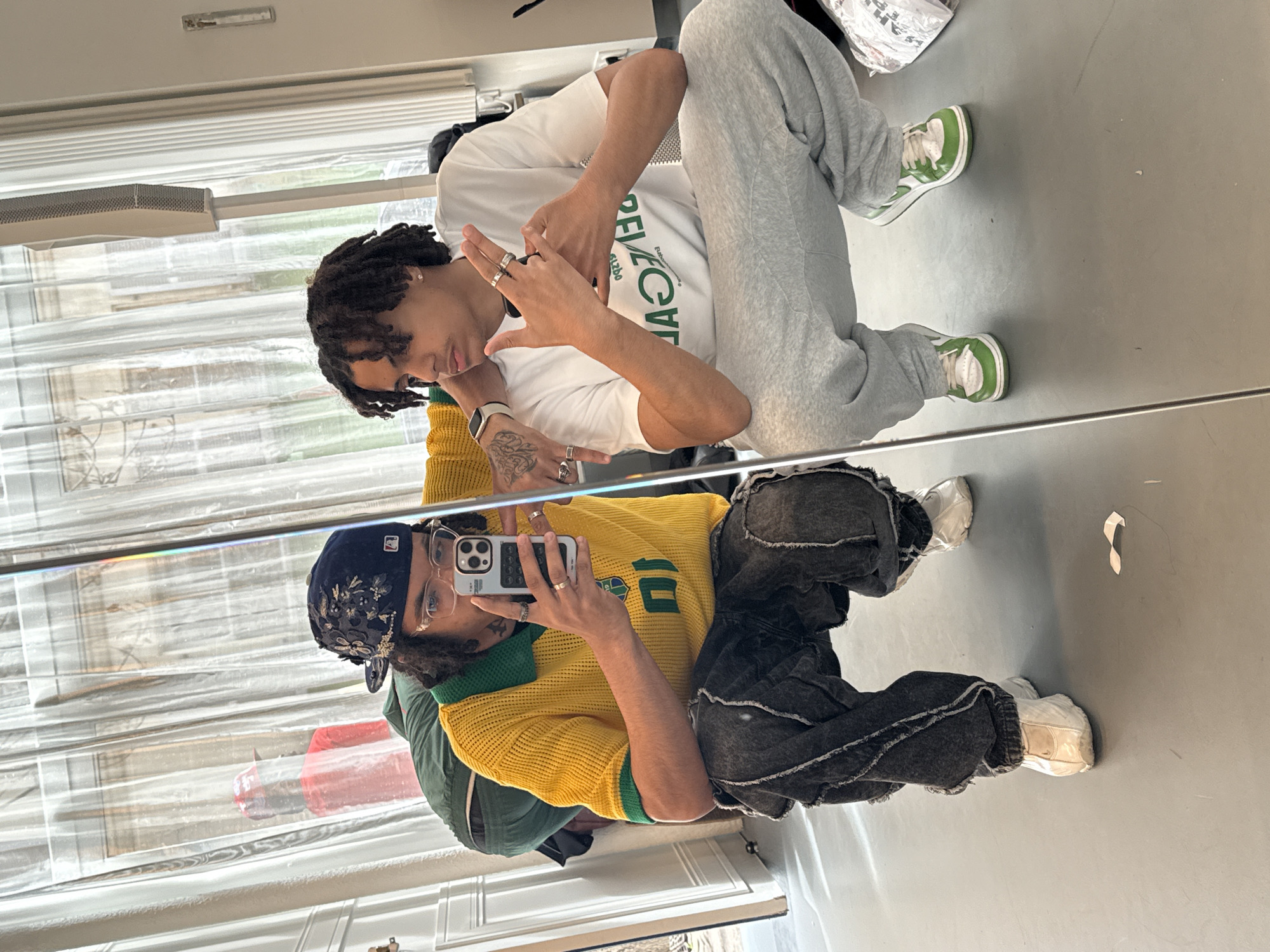 A man is posing in front of a mirror indoors, taking a selfie. He is wearing a yellow and green jersey, dark-colored distressed pants, a patterned baseball cap, and white sneakers. In the mirror's reflection is the same man, wearing a white t-shirt with green lettering, gray sweatpants, and green and white sneakers. The background appears to be an indoor space with a window. Based on the style of the window, building elements, and clothing/fashion, this image could potentially be taken in Paris, France.