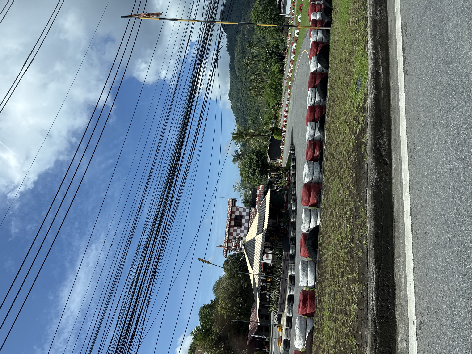 A go-kart track is visible under a bright, cloudy sky in Kathu, Thailand. The track is bordered by a row of stacked tires, painted red and white. In the background, a checkered building with a pointed roof stands next to a covered seating area. Lush green trees and a mountainous landscape fill the horizon. Numerous black electrical wires crisscross the sky, obscuring parts of the view. The scene appears to be located near a road, as indicated by the asphalt and white line in the foreground.