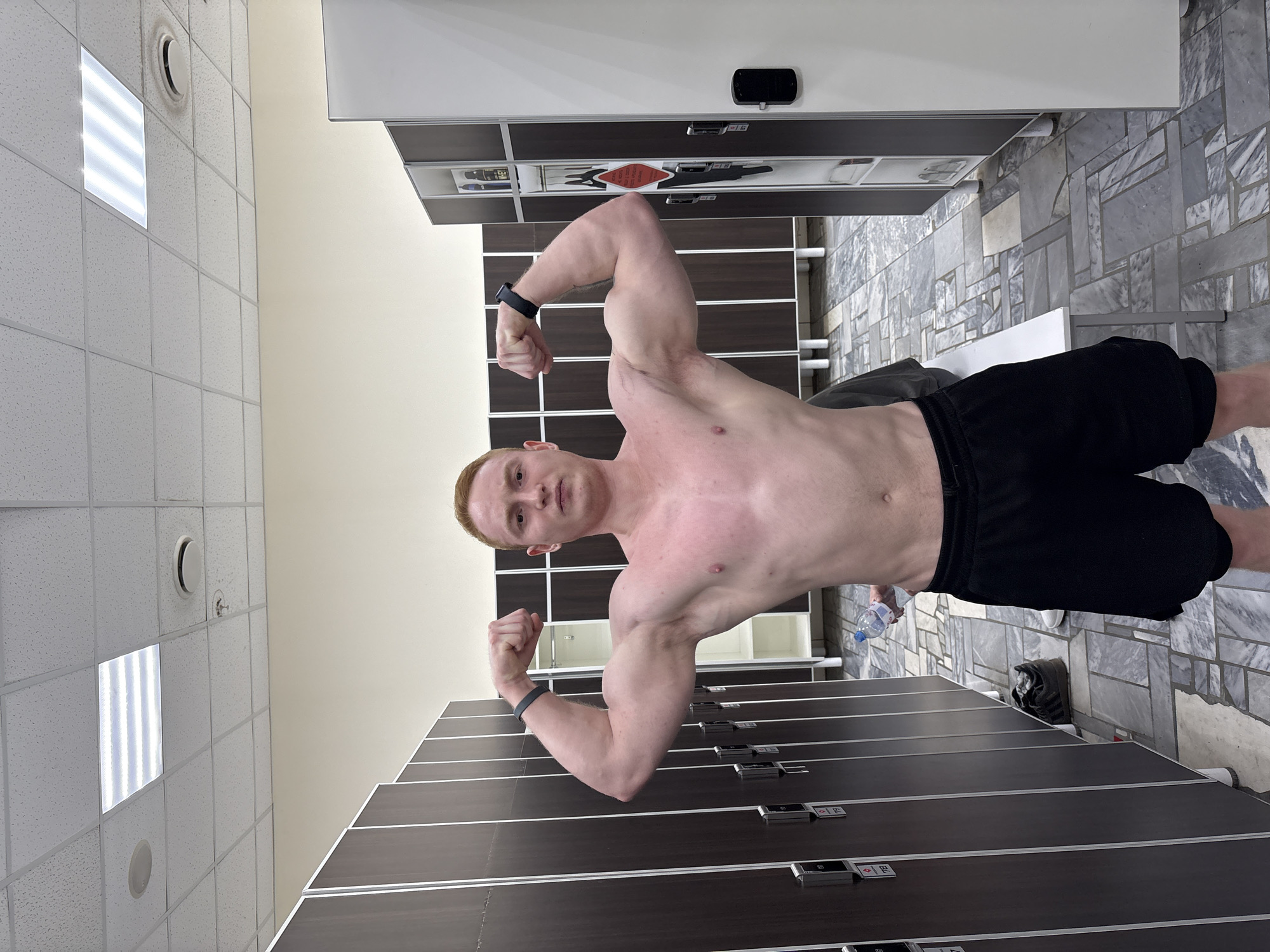 The image is a slightly rotated photograph depicting a muscular, shirtless man flexing his biceps. He is wearing black athletic shorts and a smartwatch on each wrist. His hair is light red. The man is standing in what appears to be a locker room, with rows of dark brown lockers visible behind him. The floor is tiled with grey marble-like tiles, and a white bench is partially visible in the background. On the right side of the image, a pair of black shoes and a plastic water bottle are sitting on the floor. The locker room is illuminated by ceiling lights embedded in a tiled drop ceiling. The city of Chaikovsky, Russia is not apparent in this image, and it cannot be determined from the visual information available.