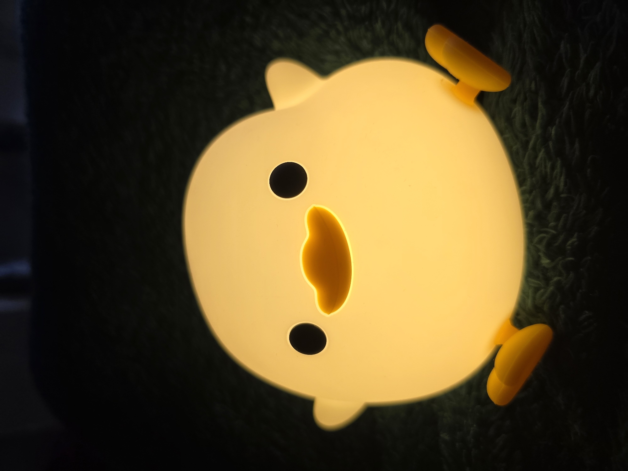 A close-up, low-angle shot shows a glowing, duck-shaped lamp resting on a dark, fuzzy surface, likely a blanket or carpet. The lamp is a soft yellow, emitting a warm light that illuminates its rounded body, two black dot eyes, and an orange beak. Two small orange feet are visible sticking out from the bottom of the lamp. The background is dark and out of focus, creating a cozy and intimate atmosphere. The scene is devoid of any human presence or activity. No specific location in Kirkel, Germany is depicted or discernible from the image.