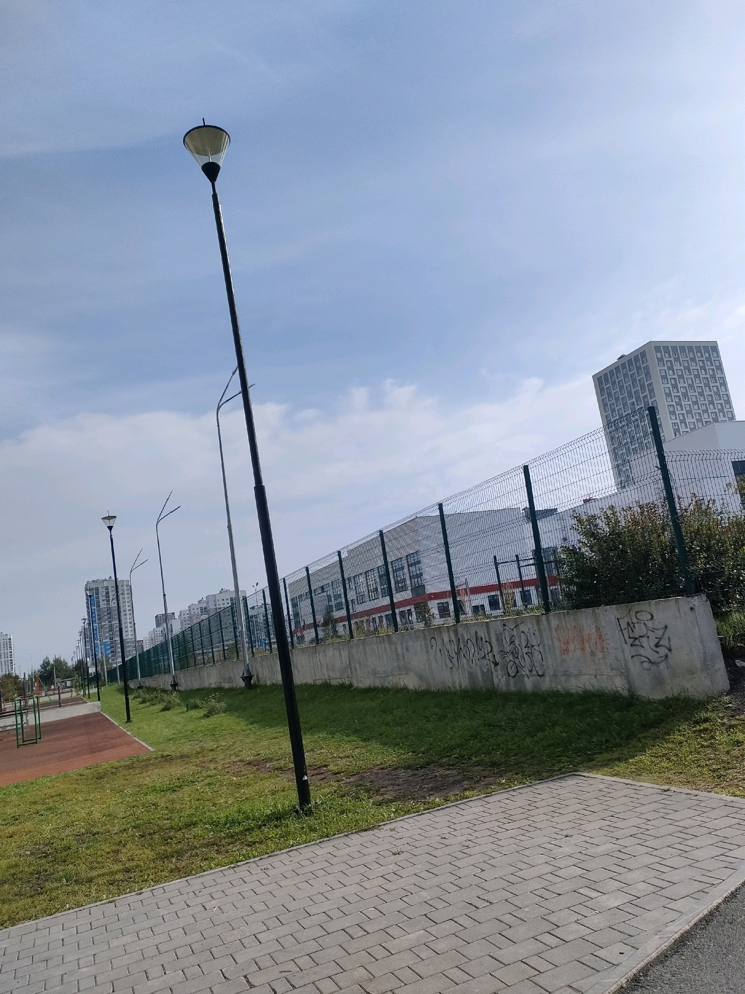 The image depicts a streetscape in Yekaterinburg, Russia. The foreground features a grey brick sidewalk. To the left of the sidewalk is a grassy area with a red-colored playground surface visible. Several tall, modern buildings with a grid-like facade are visible in the background, along with a chain-link fence. A solitary black lamppost leans at an angle in the middle ground. The sky is partly cloudy with a light blue hue.