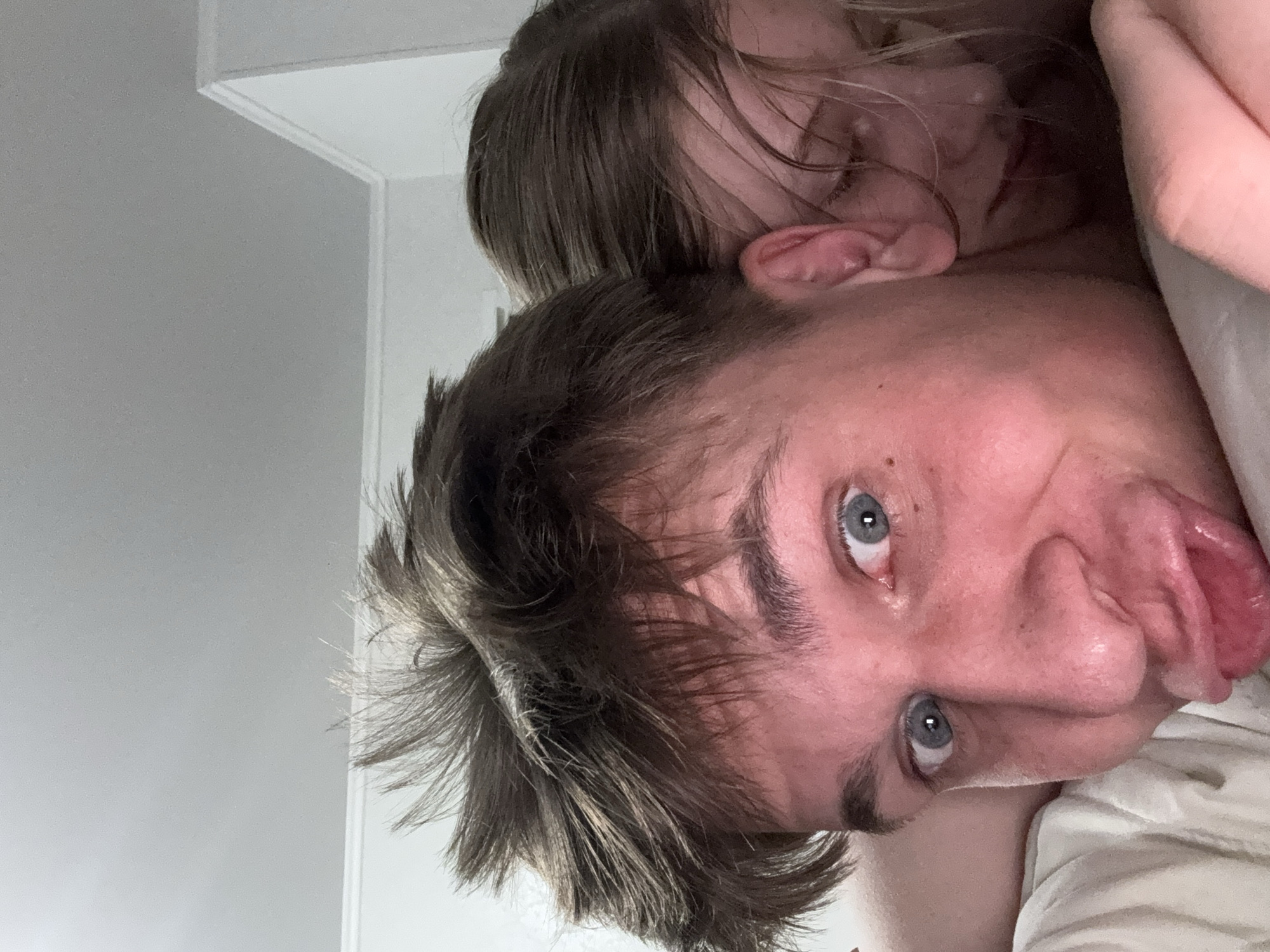A close-up selfie shows a young man with messy brown hair and blue eyes making a kissy face. He is wearing a white t-shirt. Behind him, a young woman with blonde hair is nestled against his head. The background appears to be an indoor setting, possibly a bedroom, with a plain white wall and ceiling. There are no visible elements that suggest a specific location in Tiraspol, Moldova.