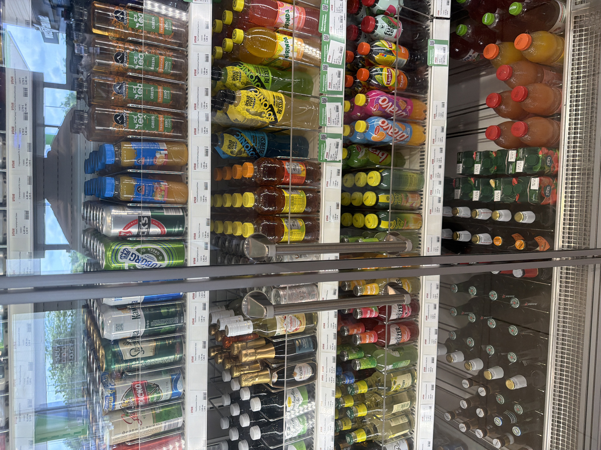 This media file depicts a brightly lit, glass-fronted refrigerated display case filled with a wide variety of beverages, likely within a supermarket or convenience store in Borovo, Bulgaria. The reflection in the glass suggests it is daytime, with light from outside or large windows visible.
The display case is organized into several sections. On the top left, multiple rows of "OLYMPUS ICE tea GREEN TROPICAL ZERO SUGAR" bottles are visible, alongside other bottled juices or drinks. Below these are shelves predominantly stocked with canned beers, including brands such as "TUBORG GREEN," "BECK'S," "STELLA ARTOIS," "STAROPRAMEN," "Kamenitza," and "Burgasko." The bottom left features glass bottles, including "MASCHIO ROSE EXTRA" sparkling wine and other bottled drinks.
On the right side of the case, a diverse selection of bottled soft drinks and juices is arranged. These include "MIRINDA," "AQUA VIVA SOTONIC RESTART," "AQUA VIVA SOTONIC REFRESH," and "AQUA VIVA SOTONIC REBOOT." Other prominent brands include "FUZE tea," "Lipton PEACH," and various "Bravo" juices. Further down, there are larger bottles, possibly from the "DEVIN" brand, and boxed "BBB" juices. The lowest shelves on the right hold smaller bottles of beverages like "BREEZER," "WKD," and "Happy Day," along with other bottled beers, some identifiable by "Heineken" and "Tuborg" branding.
Numerous price labels are visible throughout the display, predominantly displaying prices in both Bulgarian Lev (лв) and Euro (€), such as "4,29 лв / 2,19 €" and "3,69 лв / 1,89 €," indicating dual pricing. Other visible text includes product descriptions like "ELECTROLYT," "GREYPFURT" (ГРЕЙПФРУТ), and "STILSTAND ER GREEN" on a Tuborg can. No people are visible in the image; the focus is solely on the merchandise.