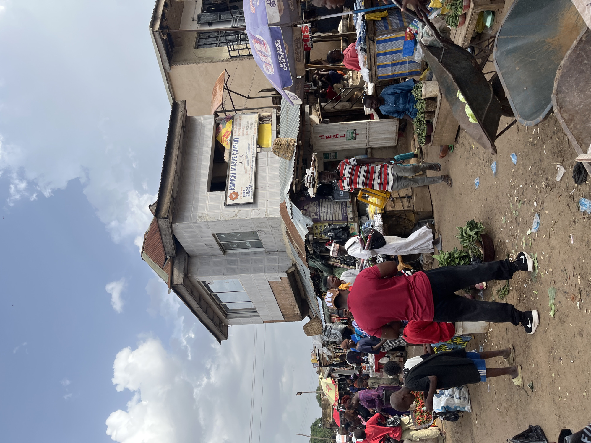 This media file captures a lively scene at Rafin Par market in Nigeria. The image is oriented vertically, presenting a bustling outdoor marketplace under a cloudy sky, suggesting it's daytime.
The primary subjects are the numerous people populating the market, engaged in various activities. Many are seen walking through the stalls, interacting with vendors, or carrying goods. The market is filled with stalls displaying a variety of items, including what appear to be fresh produce like vegetables and fruits, as well as other merchandise. Several wheeled carts, likely used for transporting goods, are visible, along with umbrellas and awnings providing shade.
Notable details include signage on the buildings, such as "AMINCHI ADASHE COMPANY" and "POS AVAILABLE HERE," indicating commercial activity and services. Text on a blue umbrella reads "Sooo Creamy Coooo." The ground is a dirt path, typical of many open-air markets, with scattered debris. The overall atmosphere is one of vibrant commerce and community interaction, with a sense of everyday market life. The presence of various individuals, including what appears to be a child in the foreground, adds to the depiction of a busy public space.