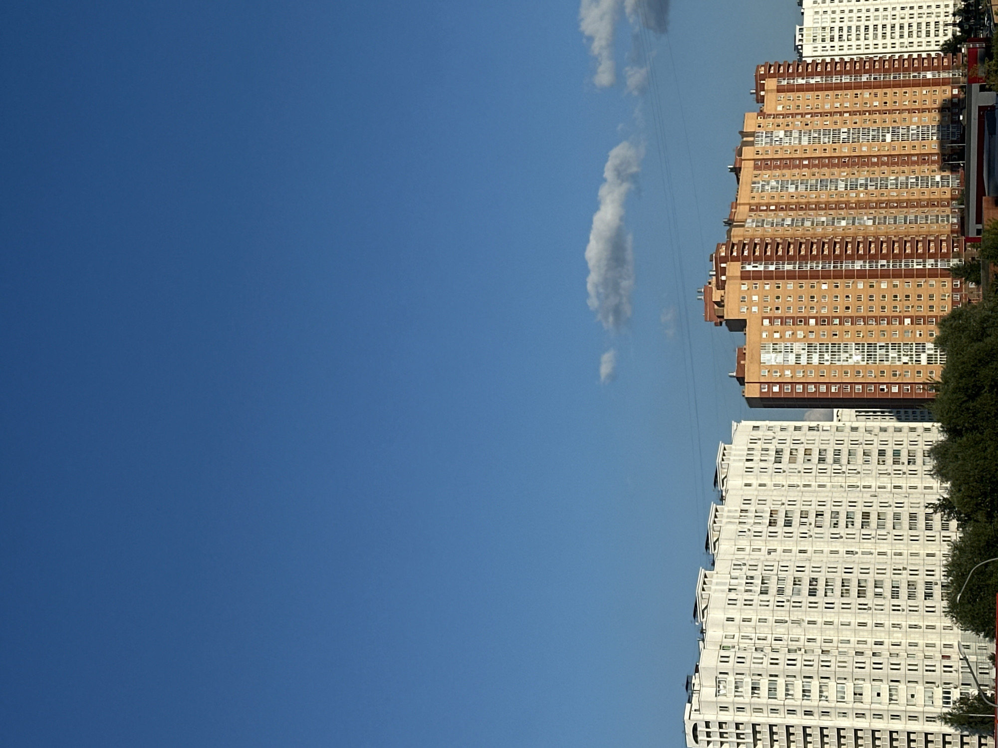The image shows a view of residential buildings in the city of Podolsk, Russia. In the foreground, there are trees. Behind the trees, two large apartment buildings are visible. The building on the left is white and has many windows. The building on the right is orange and brown and also has numerous windows. The sky above is blue with a few white clouds. Power lines are visible crossing the sky in the distance. The overall scene depicts a typical urban landscape with apartment complexes under a clear sky.