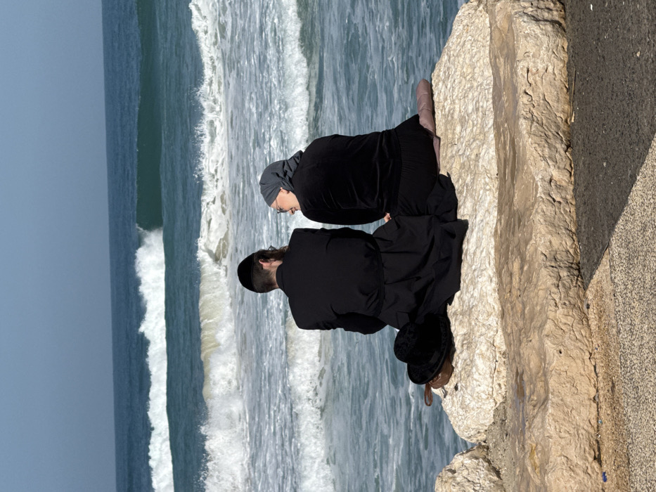 The image depicts two individuals, a male and a female, seated on large, light-colored rocks along the coastline of Tel Aviv, Israel, facing the Mediterranean Sea. The male, positioned on the left, is viewed from behind, dressed in a black suit, a long black coat, and a black yarmulke, with visible sideburns. A black felt hat and a small brown leather bag are placed on the rock immediately to his left. The female, seated to the male's right, wears dark clothing, a grey head covering, and glasses. She is turned towards the male, looking at him with an expression consistent with a smile or conversation. Both are situated with their backs to the foreground, facing the sea. The sea exhibits blue-green water with multiple breaking waves and white foam extending towards the horizon. The sky is clear and light blue. A textured paved surface is visible in the foreground, below the rocks.