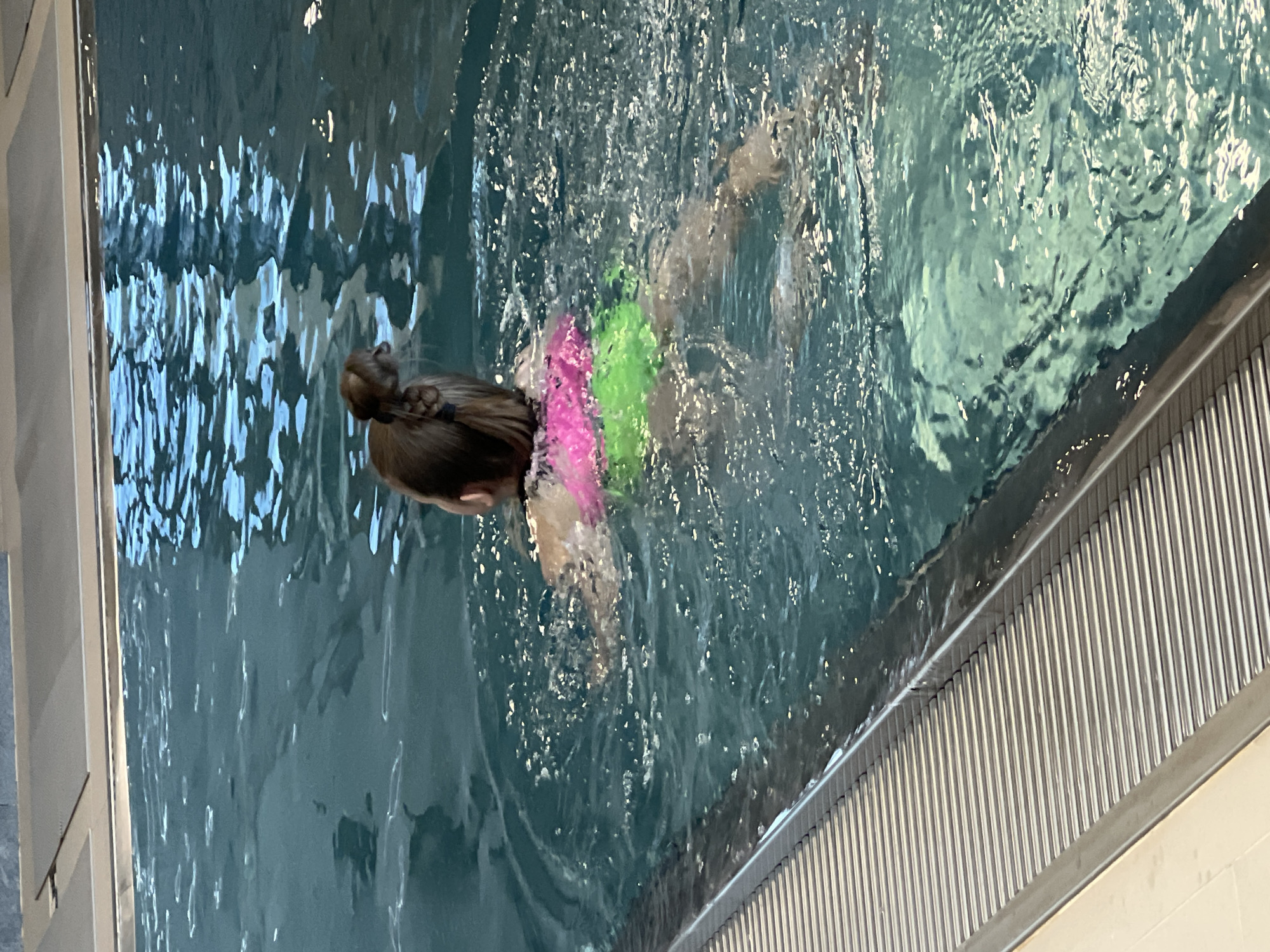 A child swims in a pool. The child is wearing flotation devices in pink and green. The pool has a metal grate on one side. The location in Chaikovsky, Russia is not evident.