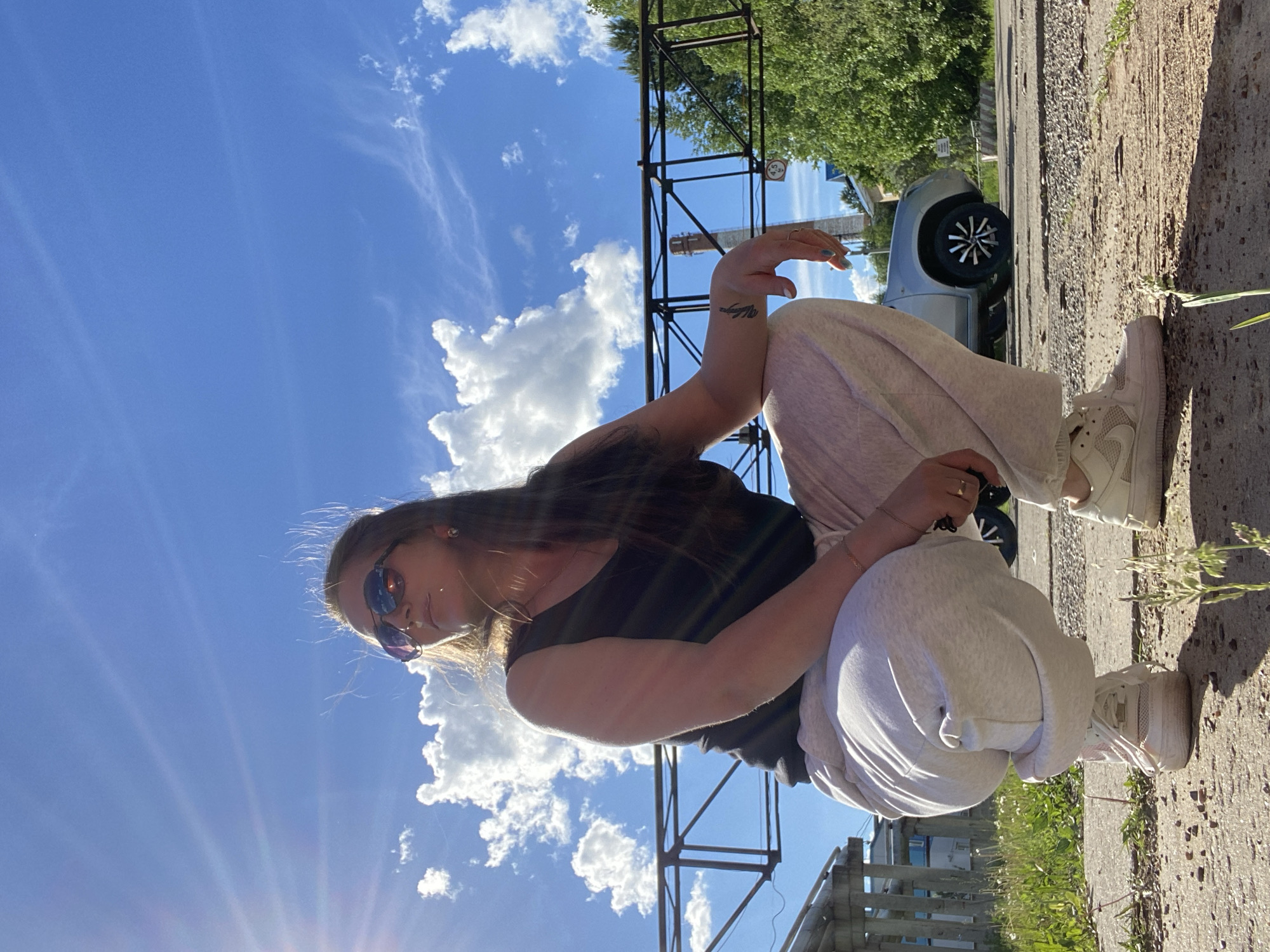 A young woman with long brown hair and sunglasses is squatting on a concrete surface on a sunny day. She is wearing a black tank top and light gray sweatpants, with white sneakers on her feet. She is looking to the side, holding what appears to be black sunglasses in her right hand. Her left arm is resting on her knee and has a tattoo on her wrist.
Behind her is a bright blue sky with fluffy white clouds, and the sun is shining from the lower left corner of the frame. The concrete surface she is on appears to be a sidewalk or a paved area.
In the background, there are elements of an urban environment, including metal construction structures, trees, and a parked car. To the right, there's a view of a street/parking area with parked cars and a fence. Based on these architectural and environmental cues, the location is most likely in the city of Chaikovsky, Russia.