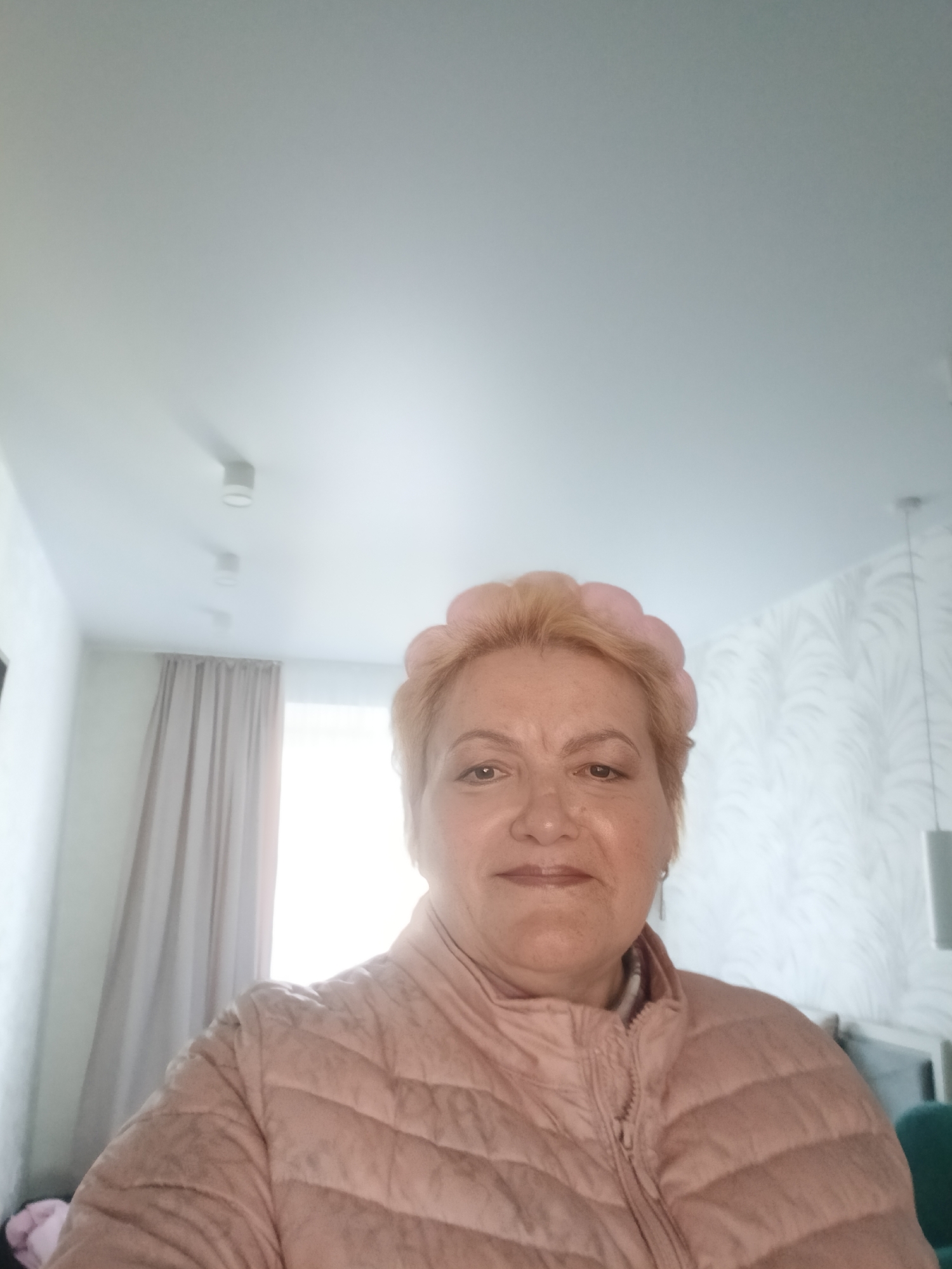 A fair-skinned woman with short blonde hair and a pink padded jacket is facing the camera. She has a pink hair accessory. The setting appears to be an interior space. To the left, a window with beige curtains is visible. In the background, there is wallpaper with a white leafy pattern. The image was taken at Murom, Russia.