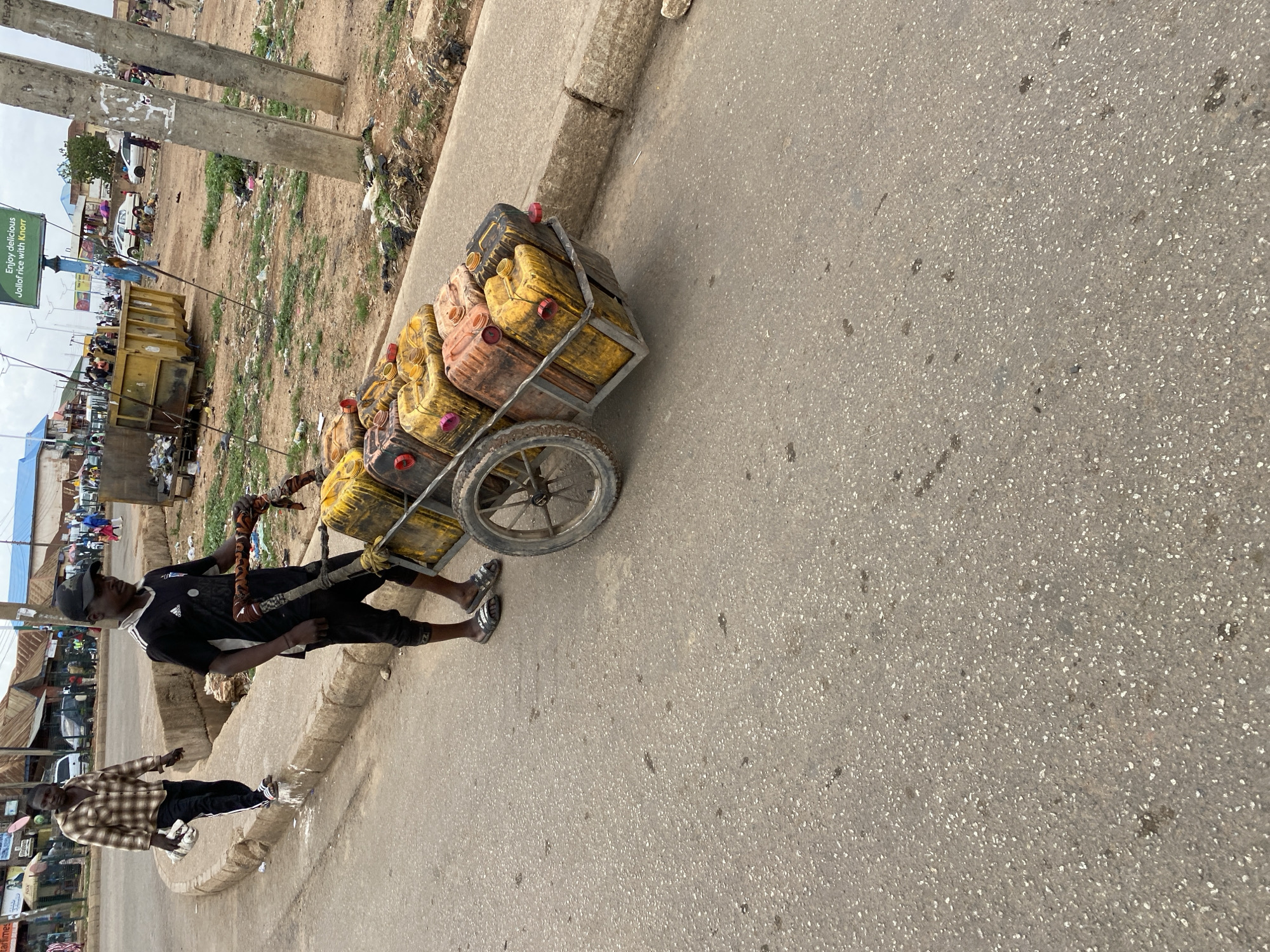 The image depicts a scene on a paved road in what appears to be Bukuru, Nigeria. A man wearing a black Adidas shirt, black pants, a black cap, and sandals is pulling a two-wheeled cart loaded with multiple yellow and orange jerrycans. Another man wearing a plaid shirt and black pants is walking alongside the cart. The background includes buildings, vehicles, and a green sign that says, “Enjoy delicious Jollof rice with Knorr.” There are areas of open dirt and debris visible adjacent to the road. The image is taken from an elevated perspective.