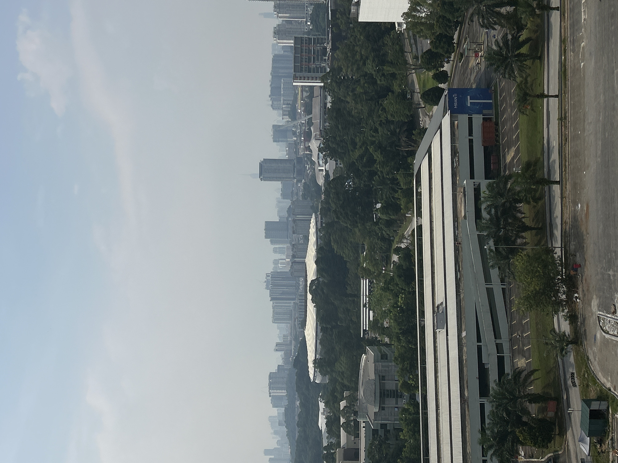 The image depicts a cityscape in Kuala Lumpur, Malaysia, under a hazy, light blue sky with scattered clouds. A dense cluster of high-rise buildings dominates the left side of the frame, fading into the distance due to atmospheric perspective. A green expanse of trees occupies the middle ground. To the right, a large, modern building with vertical white stripes stands prominently, with a blue and white "MRANTI" sign visible near the bottom of this building. A roadway runs along the right edge of the image, bordered by sidewalks and palm trees. A parking lot and other small buildings are interspersed amongst the greenery. No people are discernible in the scene.
