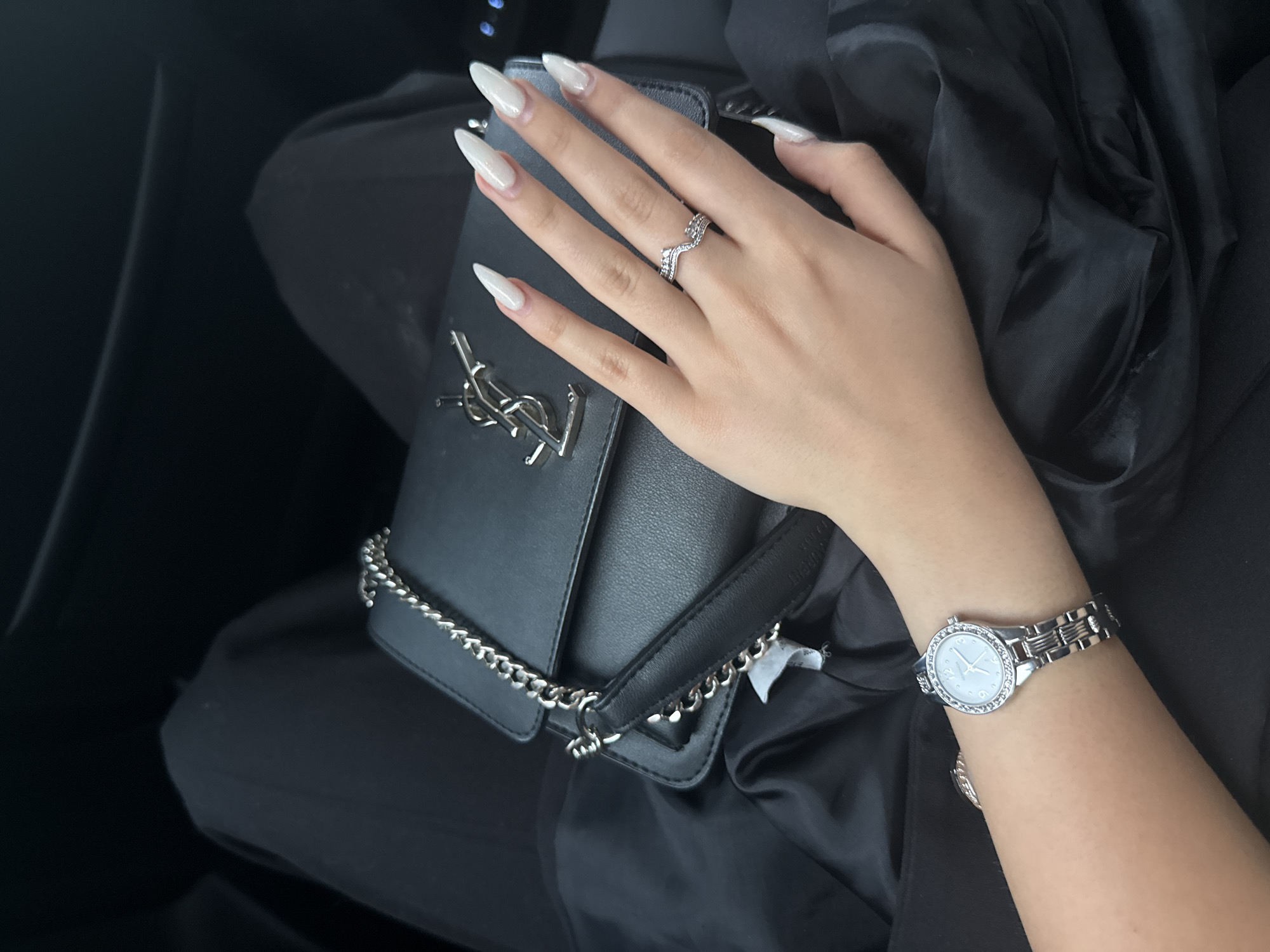 The image displays a close-up, downward-angle view of a person's upper legs, left arm, and hand, resting in what appears to be a dark interior setting. The image does not contain visual cues to determine its precise geographical location, such as the city of Sofia, Bulgaria.
The central subject is a black rectangular handbag, consistent with a shoulder or cross-body style, featuring a silver chain strap. A prominent silver YSL (Yves Saint Laurent) logo is affixed to the front flap of the bag. The bag is positioned on a surface covered with dark, smooth fabric, likely the person's lap or a seat.
A person's left hand, with a fair skin tone, is resting on the handbag. The hand displays long, pointed (stiletto or almond shape) nails, painted in a glossy, light, opaque color, possibly white or off-white. On the ring finger of the left hand, a silver-toned ring is worn, designed with a wavy or crown-like pattern and adorned with small, clear stones. On the left wrist, a silver-toned wristwatch is visible. The watch has a round face with a light, possibly mother-of-pearl or light blue, dial and a bezel encrusted with small, clear stones. The watchband is a multi-link silver-toned metal.
The person is dressed in dark, presumably black, attire; black pants are visible across the upper legs, and a section of black satin-like fabric, possibly from a jacket or blouse, is draped around the handbag and arm. The background is uniformly dark and appears to be an enclosed interior, possibly a vehicle, given the faint reflections and indistinct forms in the upper corners.