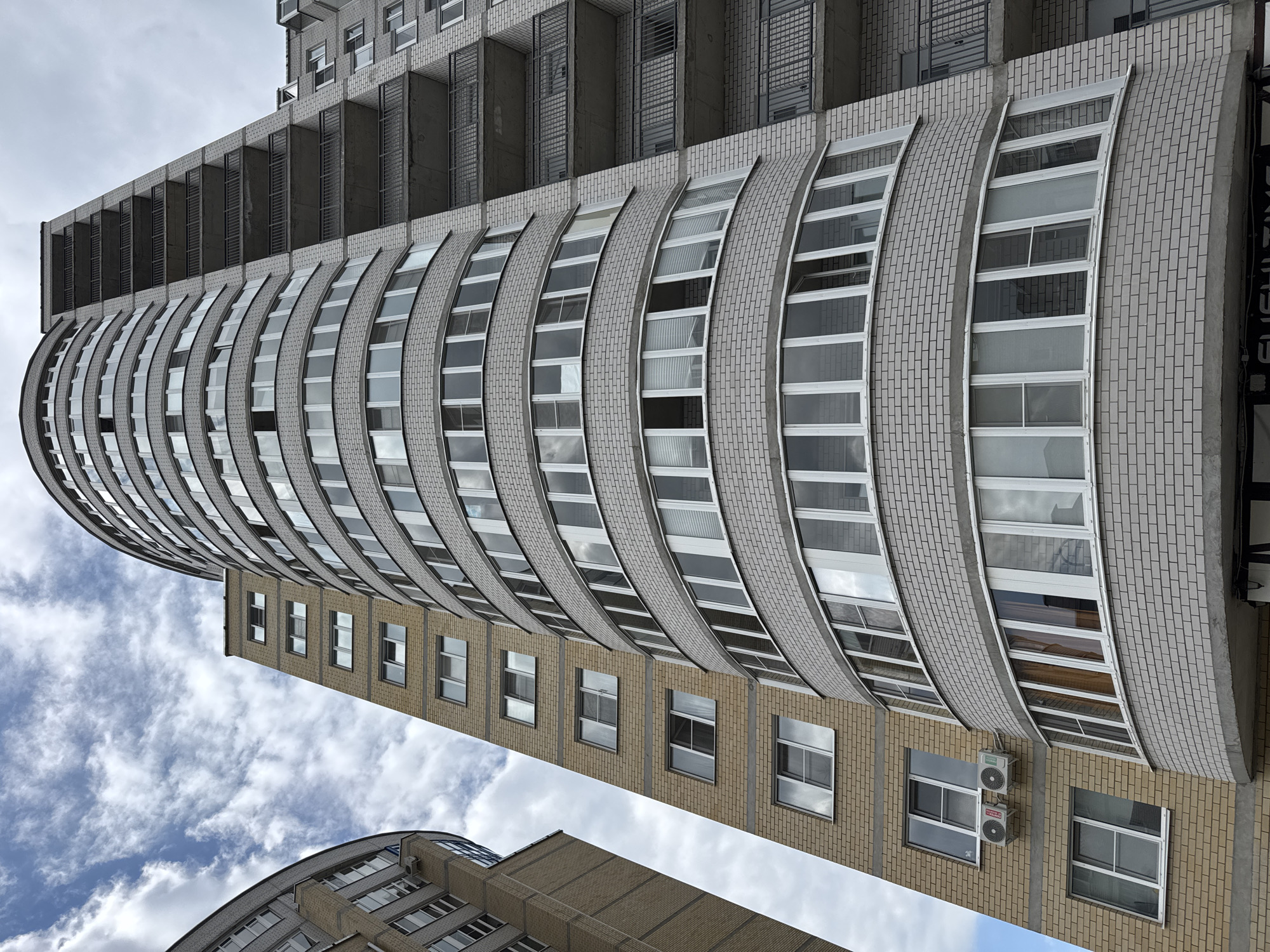 The image captures a low-angle view of a modern, multi-story apartment building in Kirishi, Russia. The building features a curved facade constructed with light grey brickwork, interspersed with numerous windows. A portion of an adjacent building, constructed with lighter colored brick and featuring a more rectilinear design, is visible on the left. The sky above is partly cloudy with visible white clouds against a light blue background. Several balconies with dark metal railings are visible on the right side of the curved building. The scene appears to be a residential area in the city.