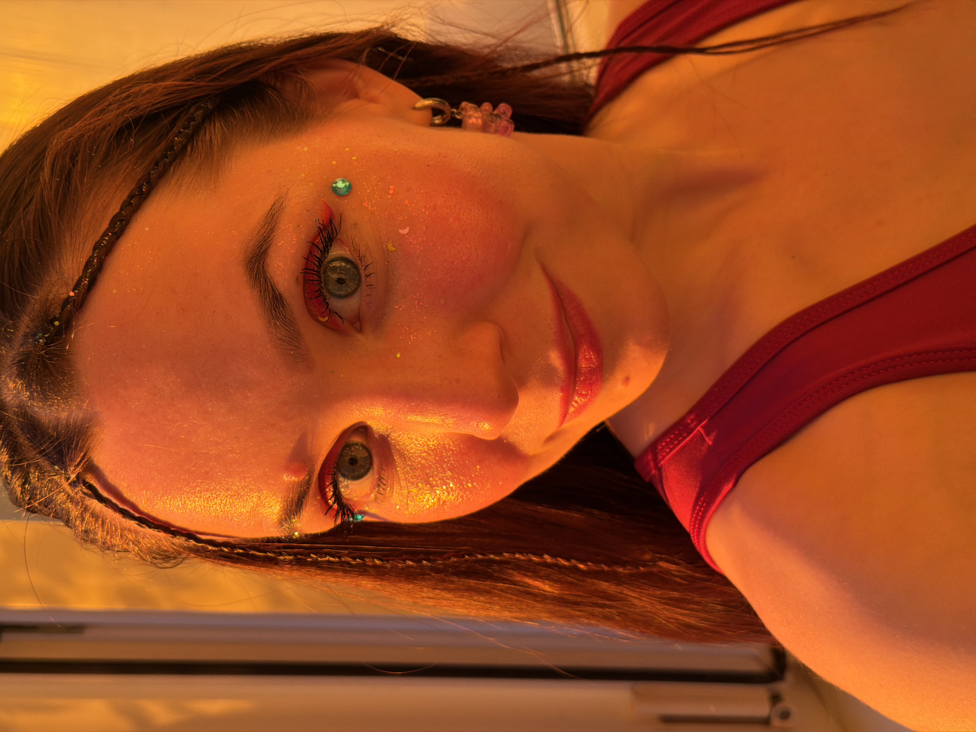 The image is a close-up portrait of a young woman with dark hair styled in braids, wearing a red top. She has striking green eyes and her face is adorned with glitter and colorful makeup. She is wearing a small hoop earring with a pink gummy bear charm. The lighting is warm and orange-toned, suggesting either sunset or artificial lighting. There is no discernible city or specific location in Chaikovsky, Russia, visible in this image.