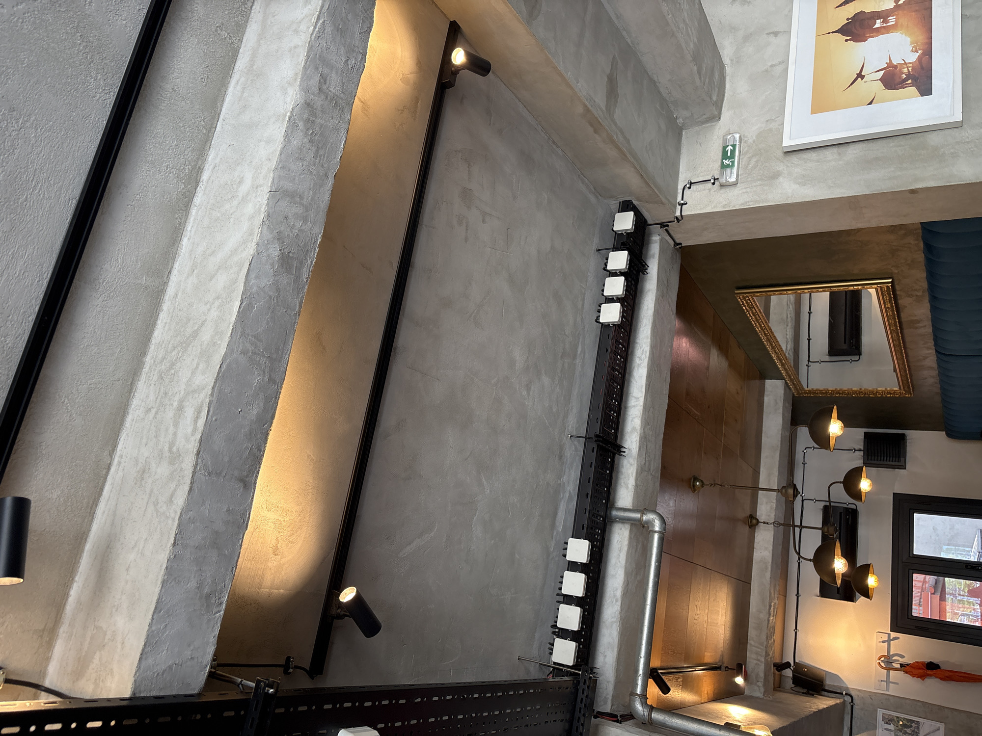 Captured from a low-angle perspective, the photograph showcases a modern interior featuring a concrete ceiling intersected by black fixtures and lighting elements. Positioned at the right of the frame, a wall adorns a framed artwork and a mirror, alongside industrial-style lighting composed of metal pipes and bulbous lamps. A window provides natural light, illuminating elements within. The composition emphasizes geometric shapes and industrial design, creating a visually striking and detailed portrayal of the interior space. The location is Unknown, Unknown.