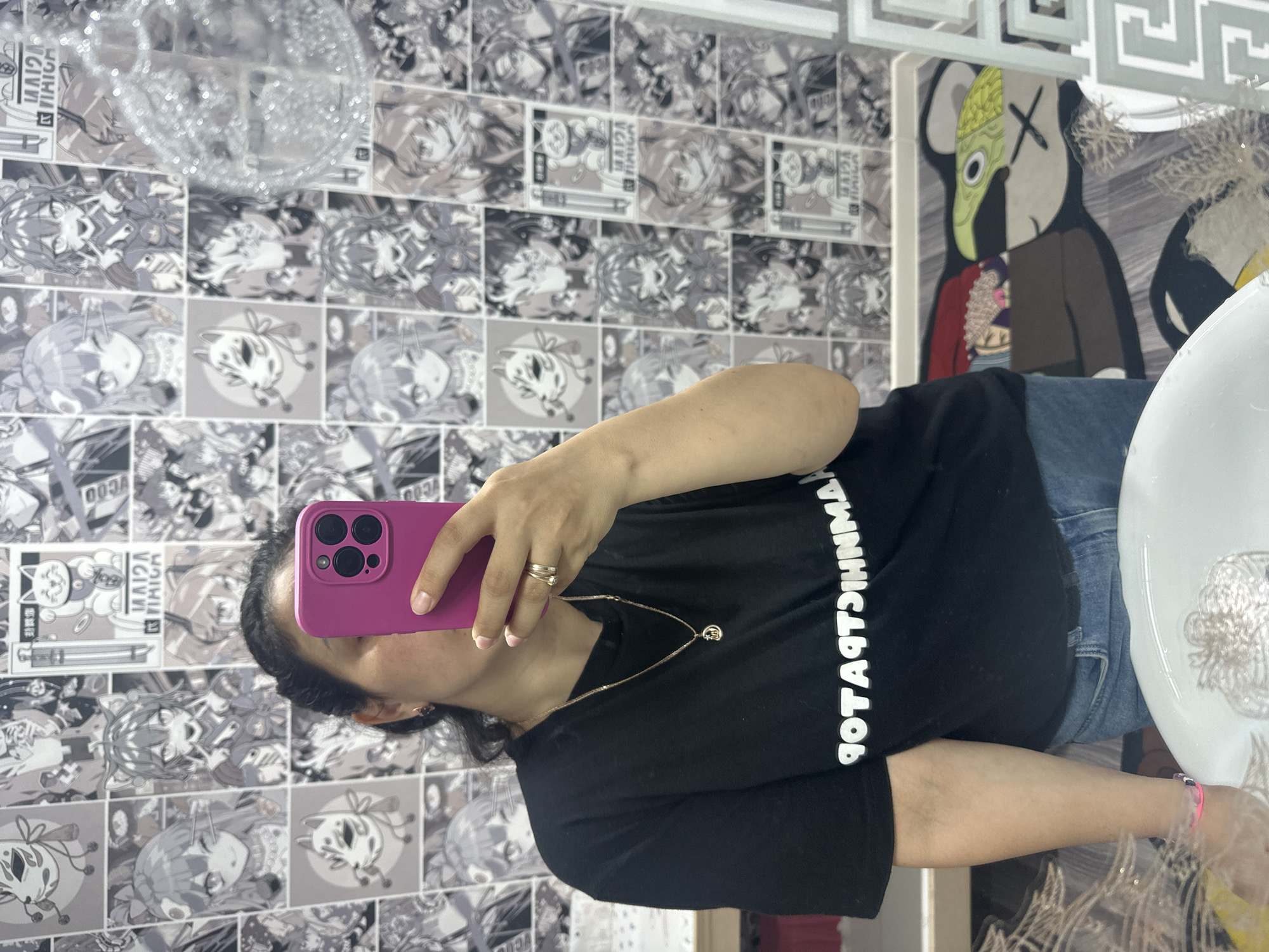 A woman is taking a photo of herself in a bathroom. She is wearing a black T-shirt and blue jeans, and she has a gold necklace. She is holding a pink smartphone up to her face. The bathroom has anime-themed wallpaper, a sink, and a KAWS figure.